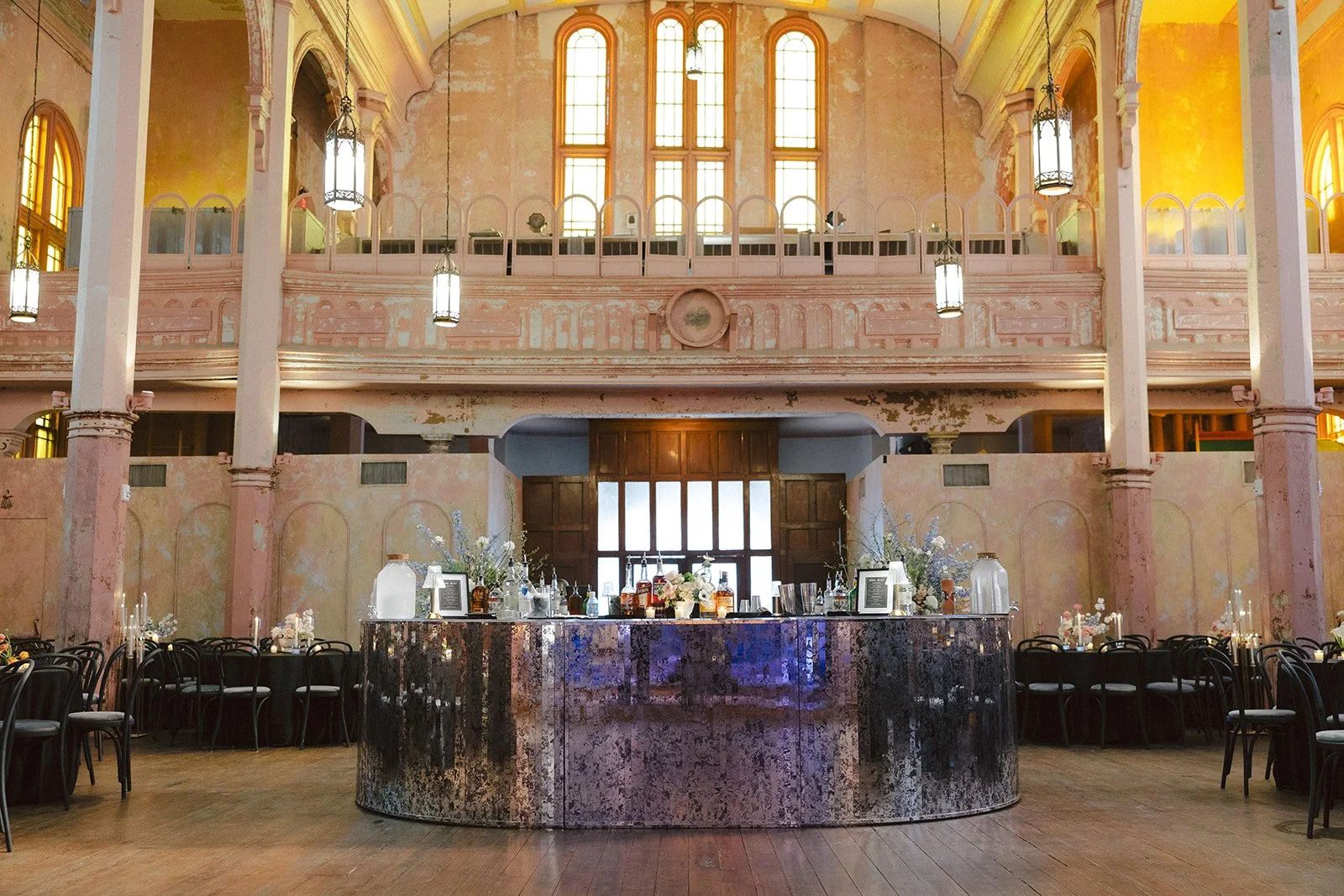 Interior of a vintage-style event hall with high arched stained glass windows, a two-story balcony, and a dark marble bar in the center decorated with flowers, candles, and glass bottles. Seating arrangements with black chairs are on either side of t