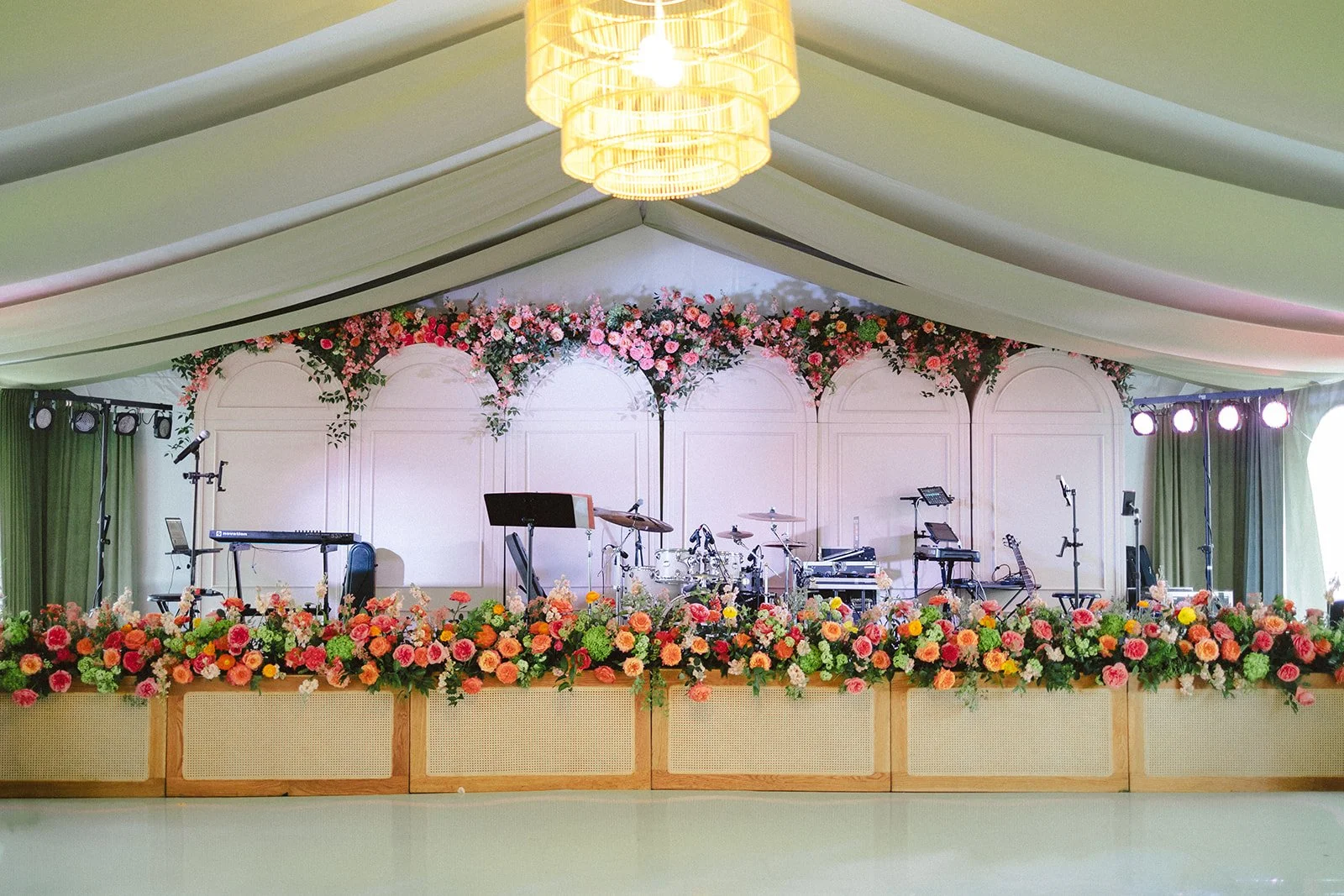 Empty wedding or event stage decorated with an abundance of pink and orange flowers, with musical instruments, microphones, and lighting equipment set up, under a large cream-colored tent ceiling.