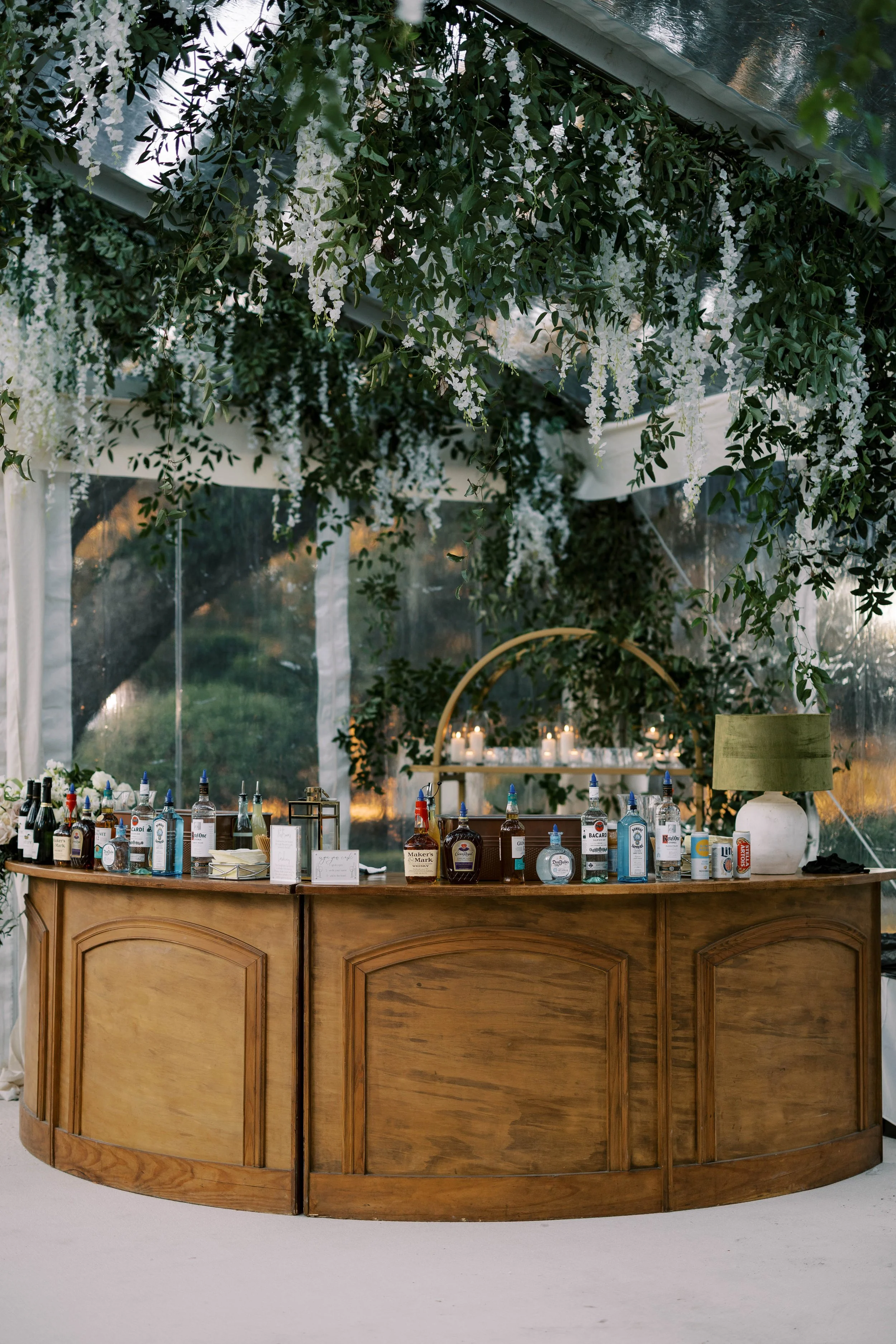 A wooden bar counter with various bottles of alcohol, a large green lamp, and a vase. Behind the bar, there are hanging white flowers and green foliage, with a transparent tent and a view of trees outside.