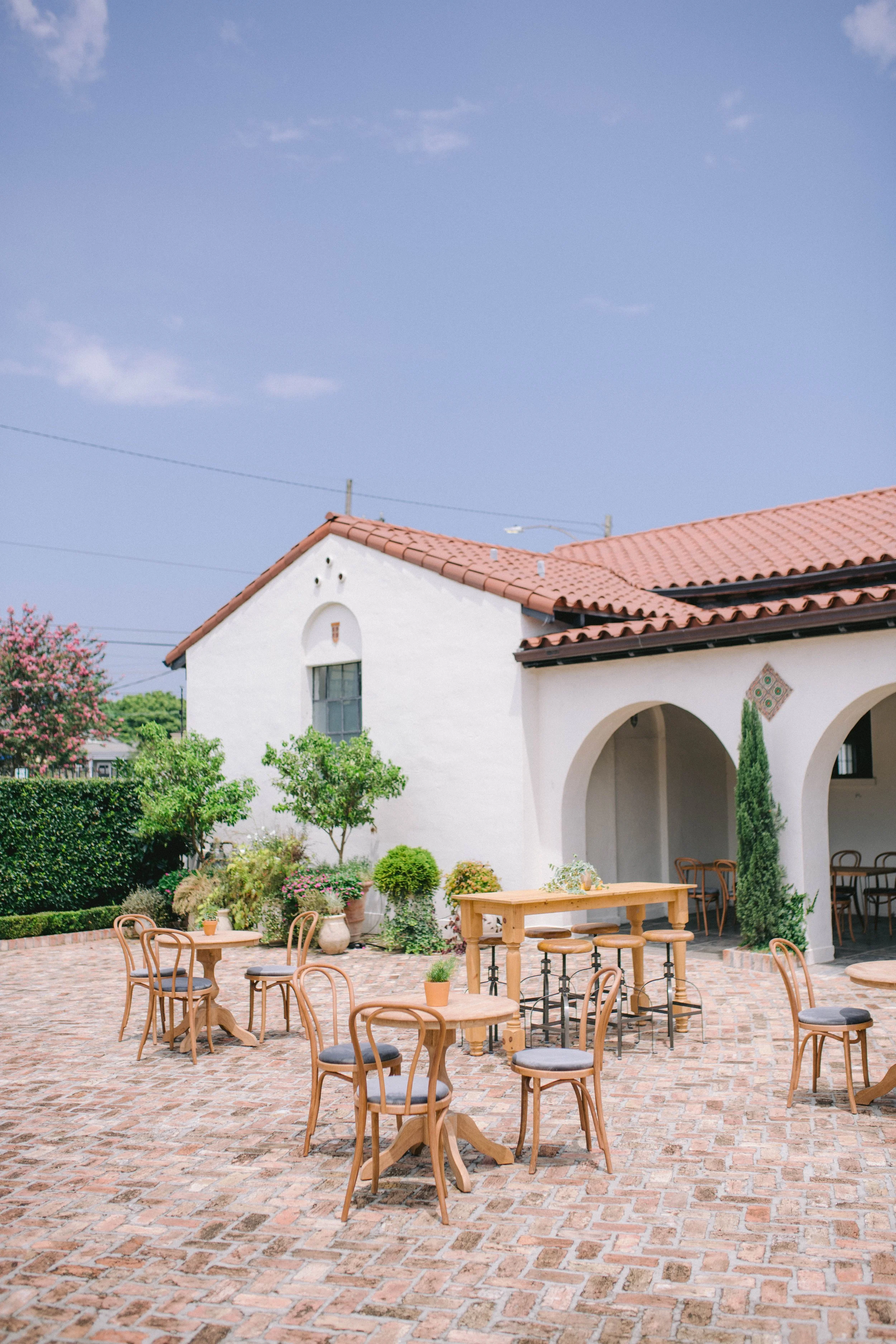 Outdoor patio with brick flooring, wooden tables and chairs, surrounded by lush green plants and trees, with a white stucco building featuring a red-tiled roof and arched entryway, under a blue sky with a few clouds.