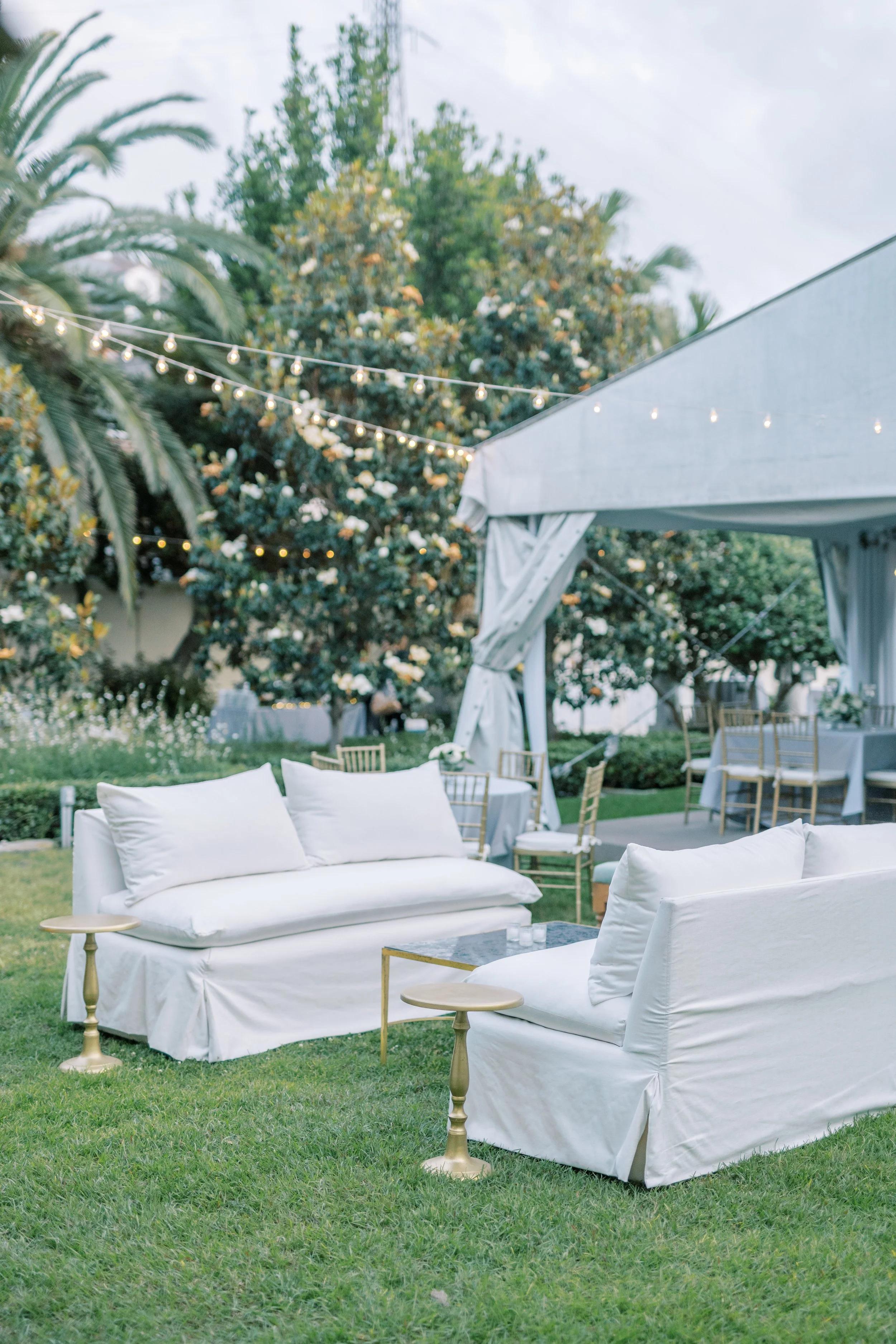 Outdoor wedding reception with white sofas, wooden tables, and string lights on a lawn with trees and cloudy sky.