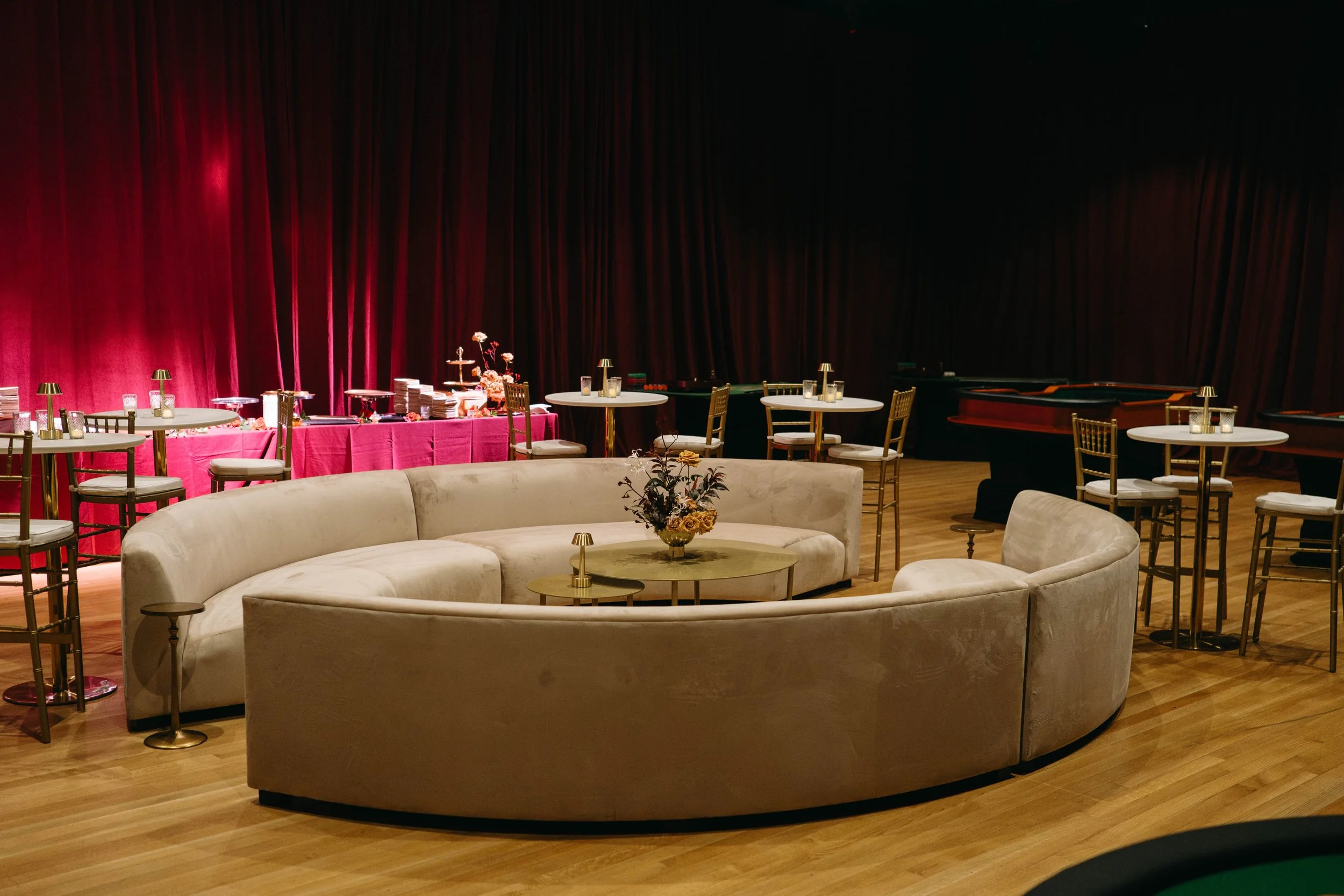 An event space with a dark red curtain backdrop, decorated tables, and a floral centerpiece, with a curved white sofa in the foreground.