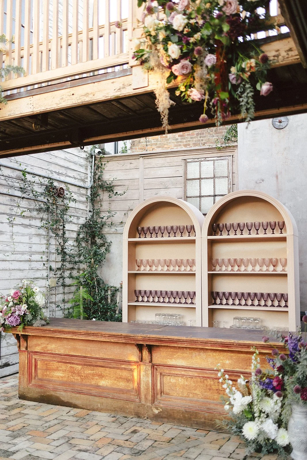 An outdoor setting with a wooden bar counter, floral arrangements on each side, a shelving unit filled with purple glasses, surrounded by brick and weathered wood walls, and a floral hanging decoration overhead.