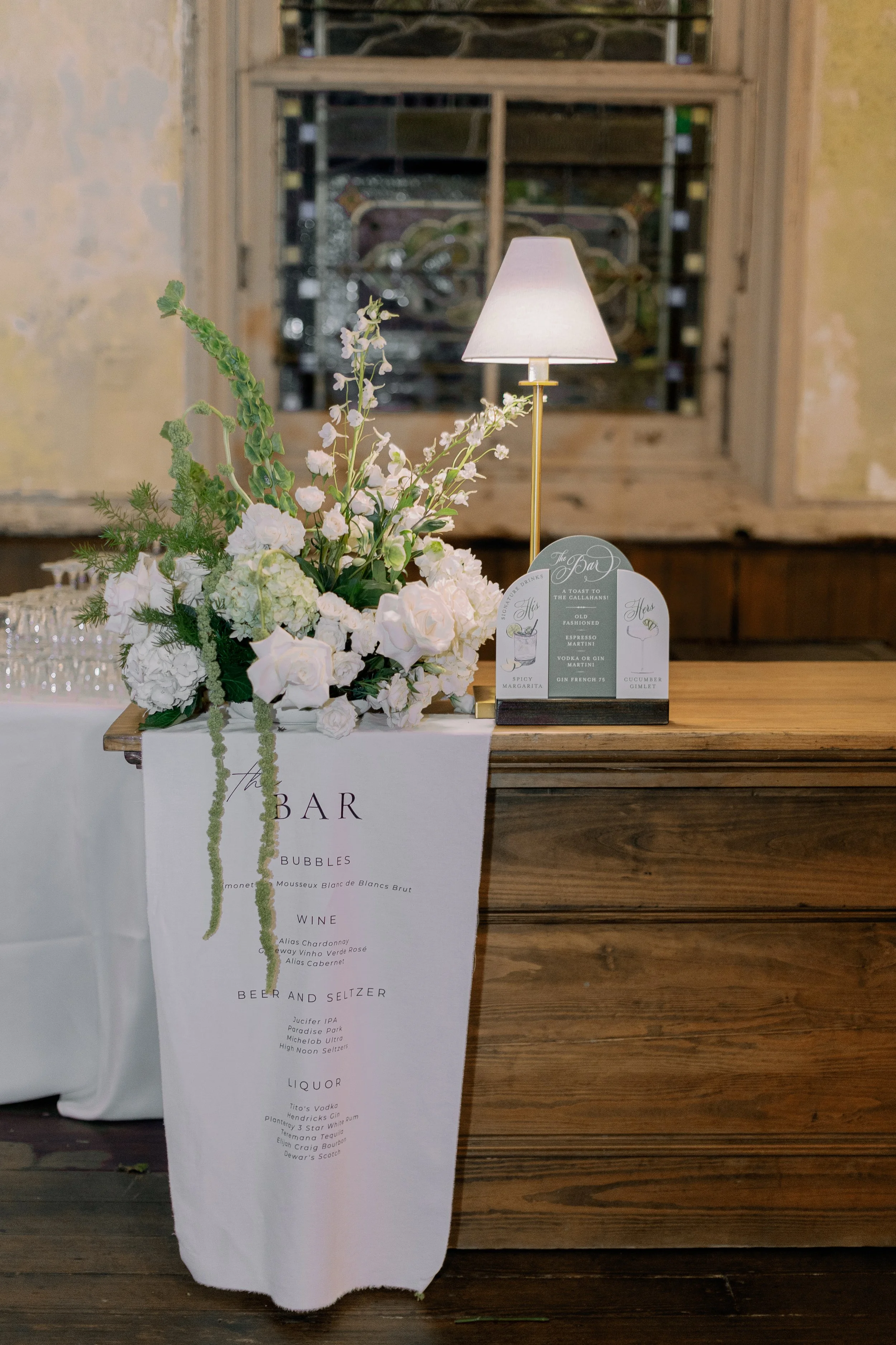 A floral arrangement with white flowers and greenery on a table, with a small lamp beside it. A menu or sign listing drinks and beverages hangs over the table.
