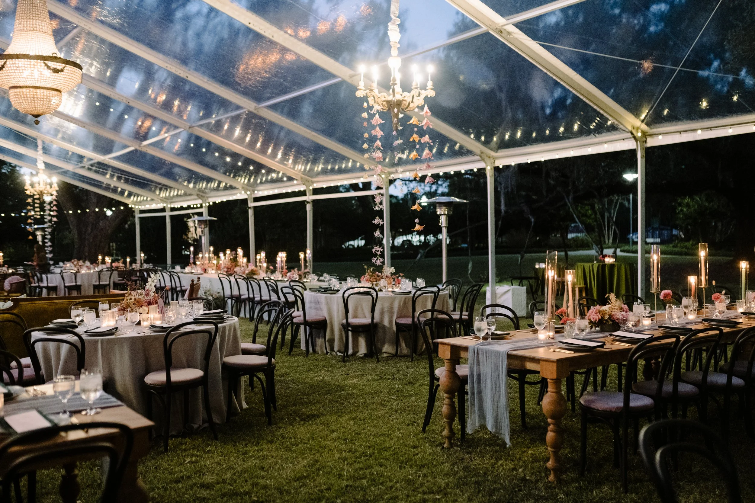 Outdoor event space set up with round tables covered in white tablecloths, adorned with floral centerpieces and lit candles. The space is under a clear tent with string lights and chandeliers, creating a warm, elegant atmosphere for evening dining.
