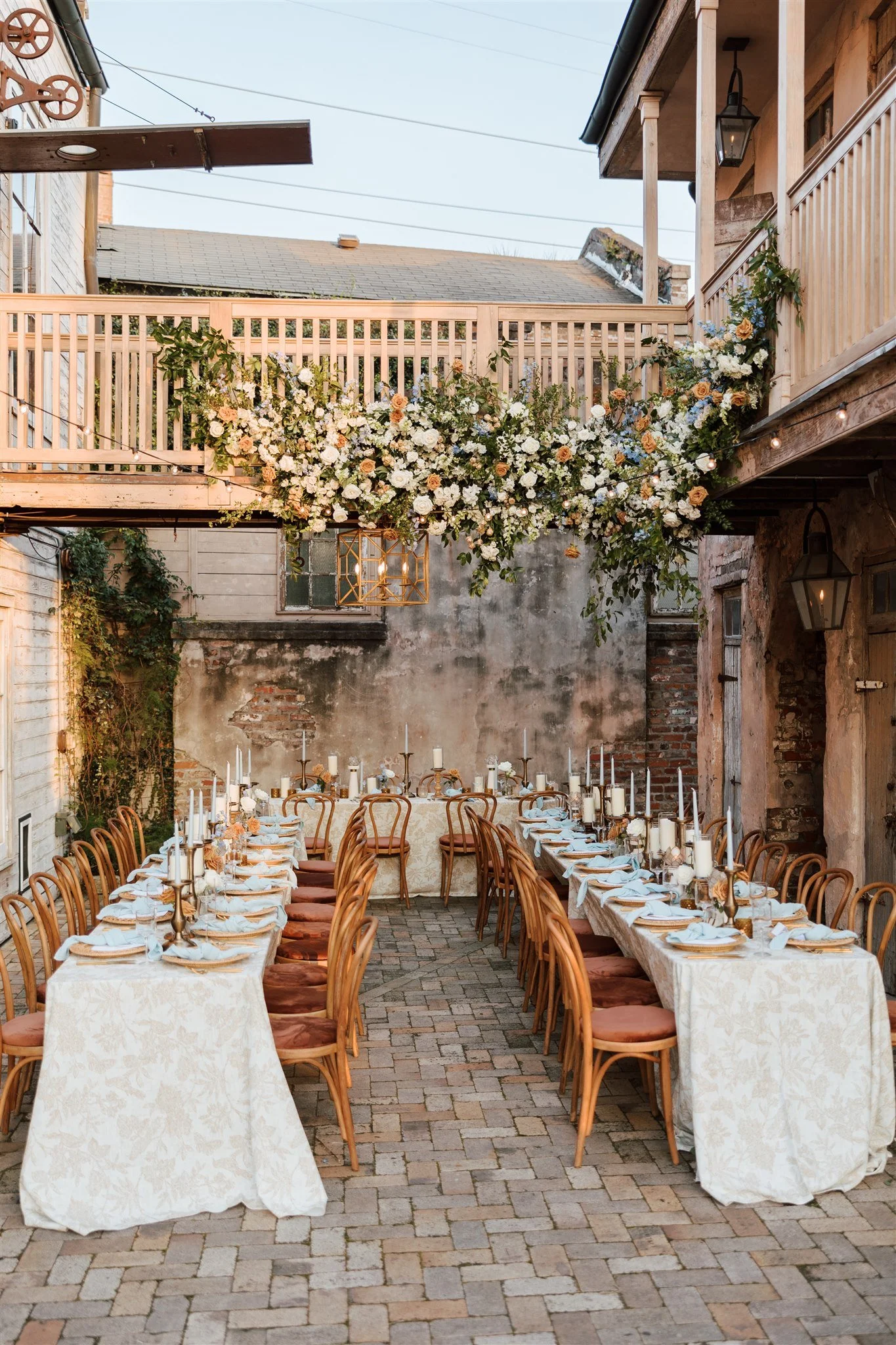 Long dining table set with white tablecloths, candles, and elegant tableware, surrounded by wooden chairs, in a rustic outdoor courtyard decorated for a celebration, with a floral arrangement hanging from the balcony above.