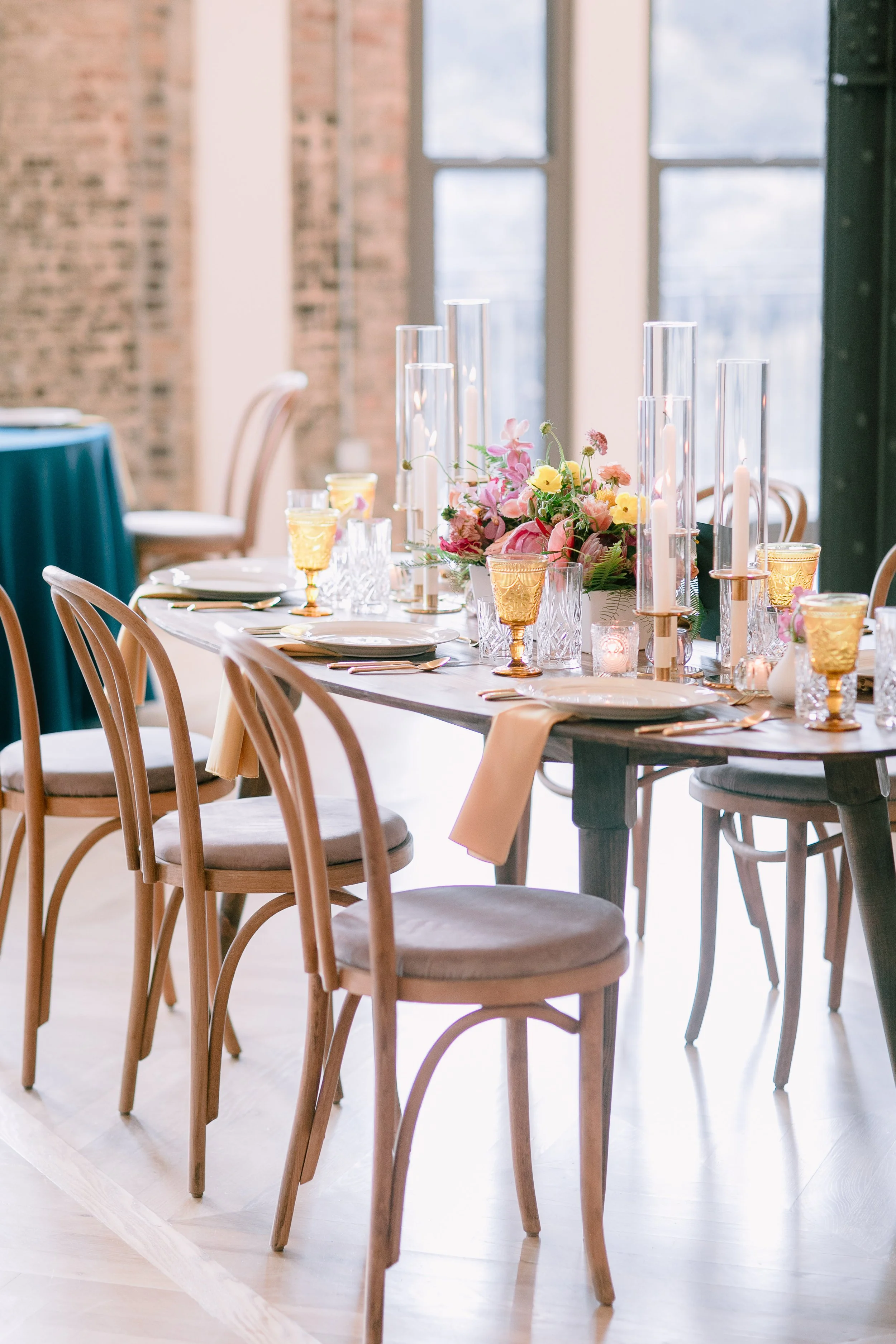 A dining table set with pink and yellow floral centerpiece, candles, glassware, and gold-colored cups in a bright room with large windows, brick walls, and wooden chairs.