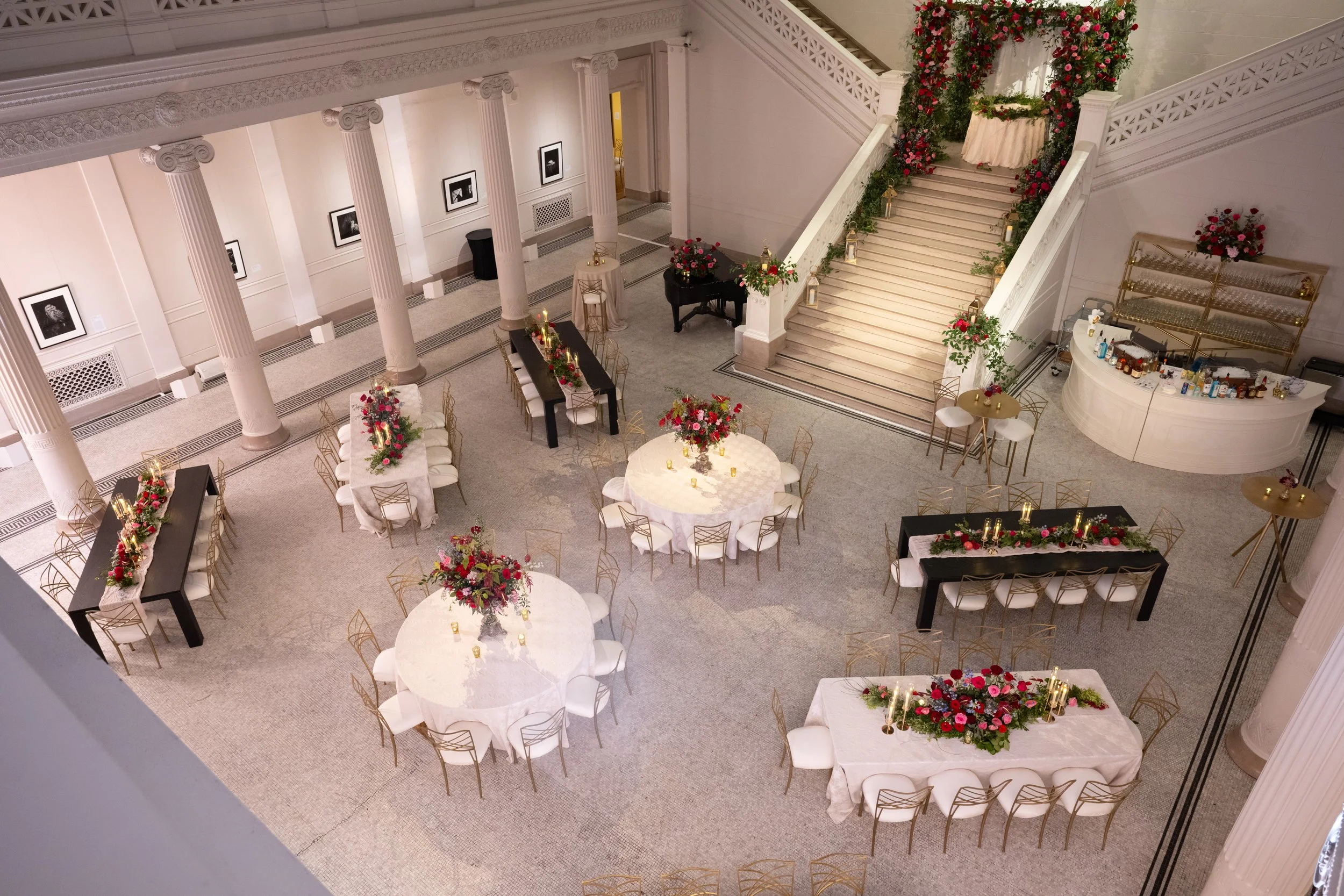 Elegant decorated event space with round tables and floral centerpieces, a staircase with floral decorations, and a bar area with bottles, all in a grand interior with white walls and columns.