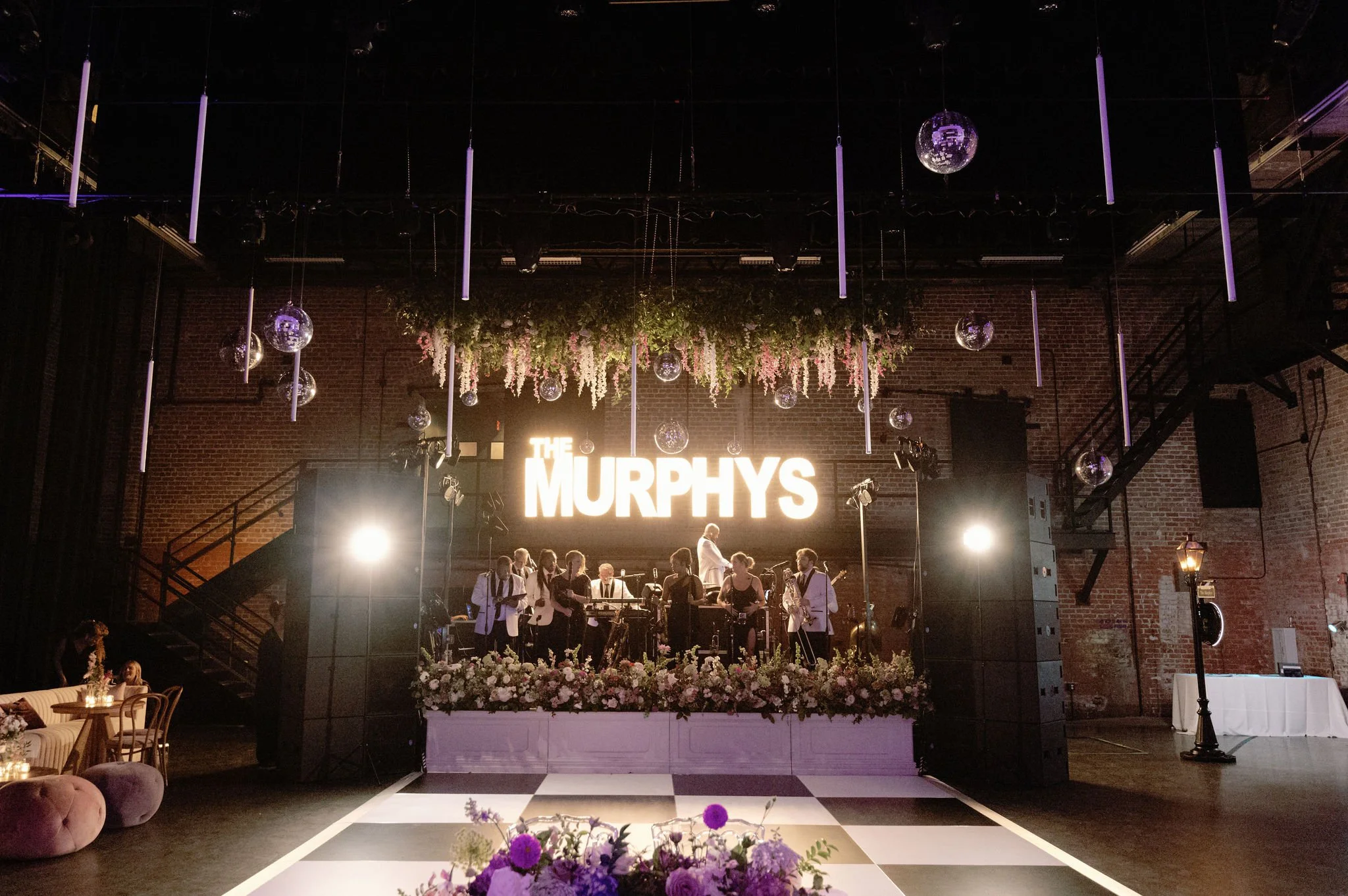 Indoor event space with live band and floral decorations, large illuminated sign reading 'THE MURPHYS', hanging disco balls and purple lighting, with tables and seating.
