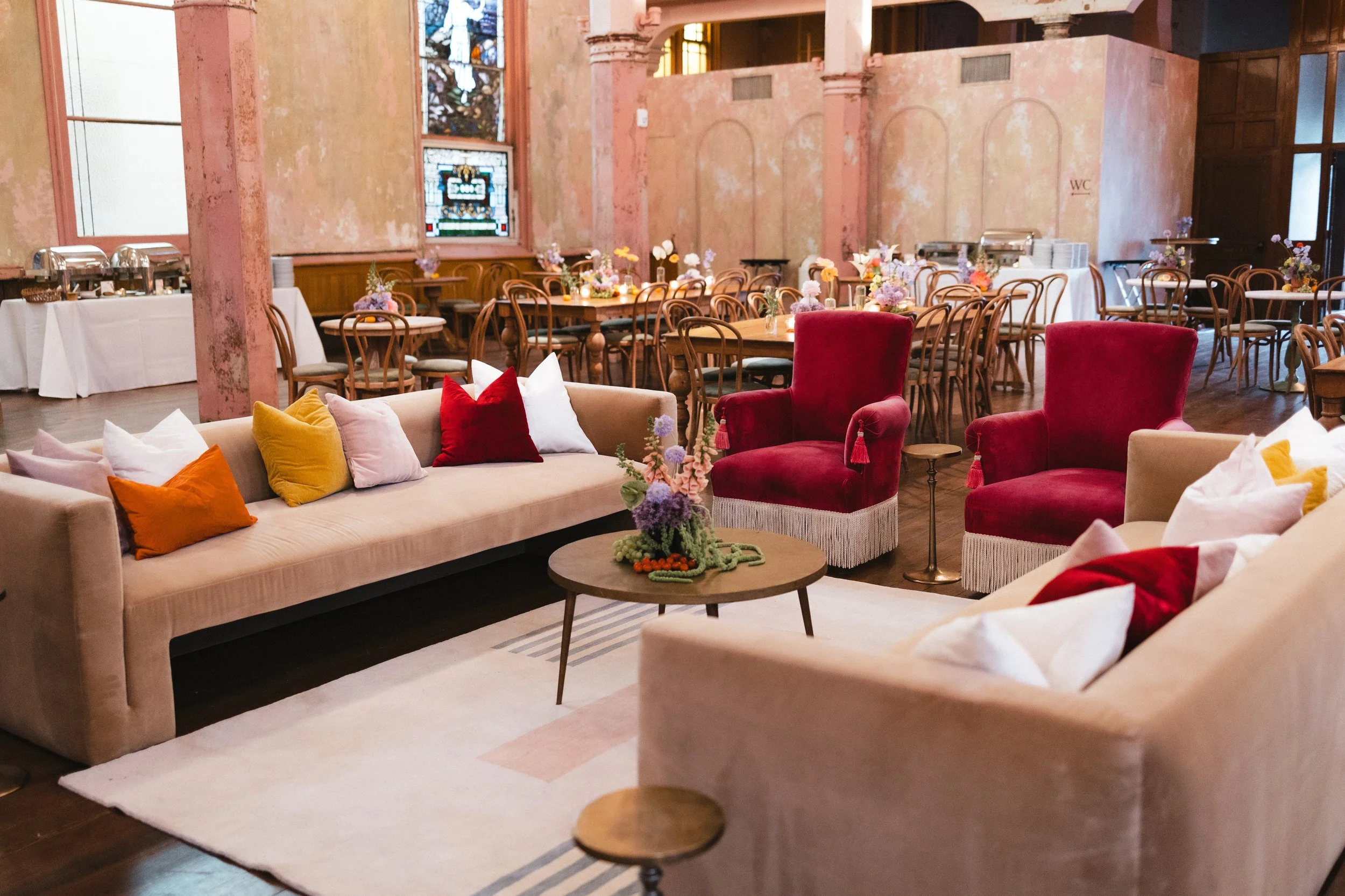 Elegant event space with plush beige and red velvet chairs, a beige sofa with colorful pillows, and decorated tables with flower arrangements; stained glass windows and distressed pink walls in the background.