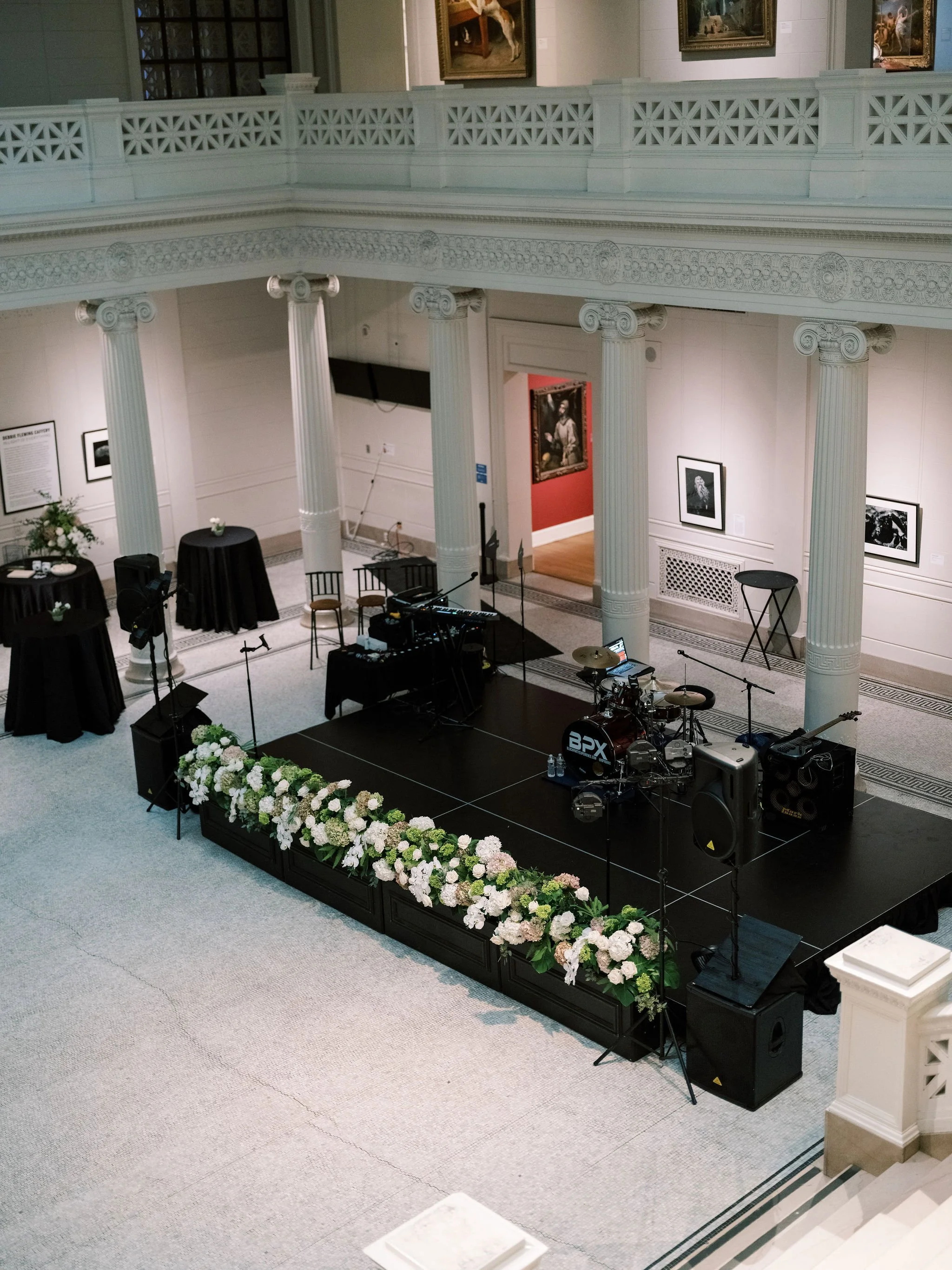 An indoor event space with a stage decorated with a floral arrangement. The stage has musical instruments, microphones, and sound equipment set up. The hall features large white columns, ornate molding, and framed artwork on the walls.