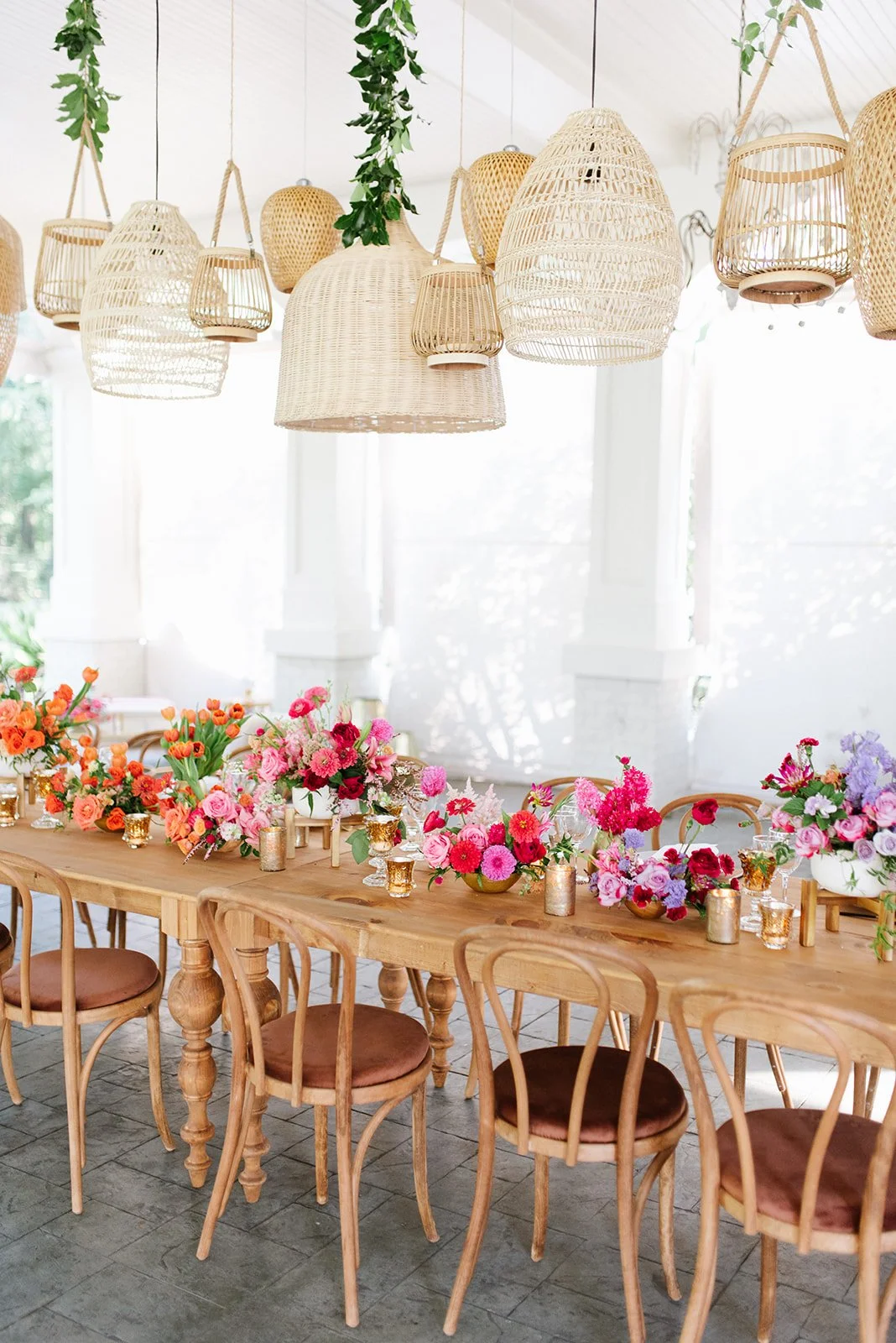 A dining table decorated with colorful flowers, gold candles, and glassware in a bright, airy room with rattan pendant lights hanging from the ceiling.