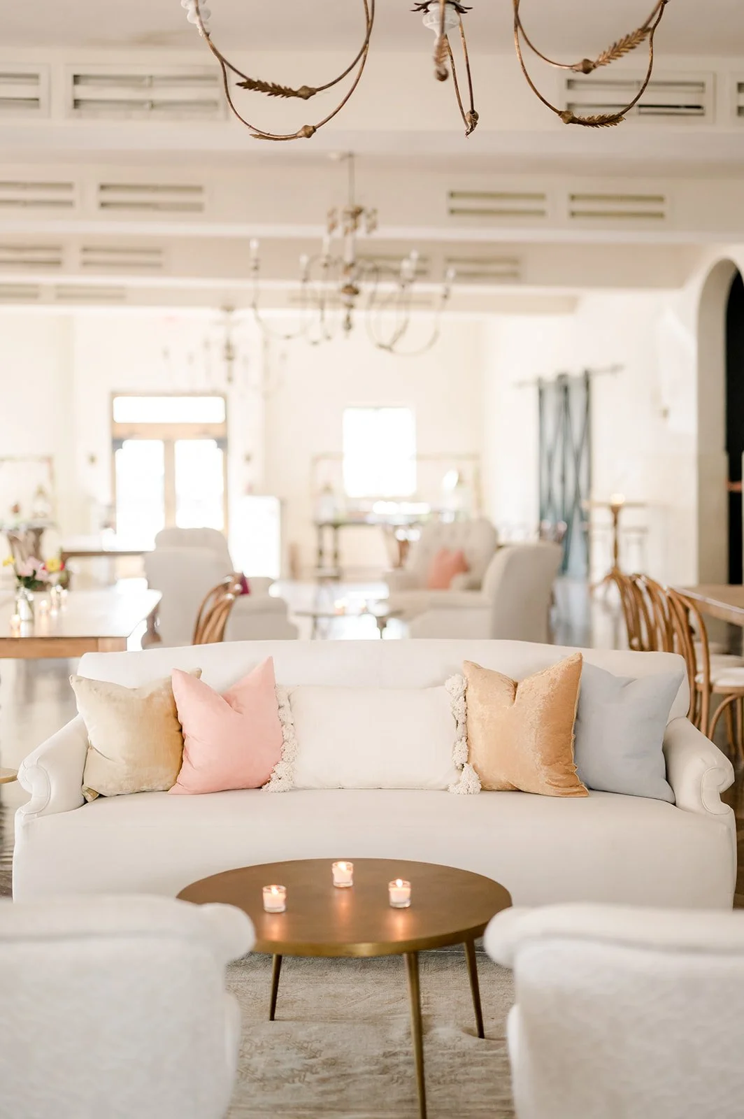 A bright, airy living room with a white sofa decorated with pastel pillows, a round wooden coffee table with candles, and multiple chandeliers hanging from a white ceiling.
