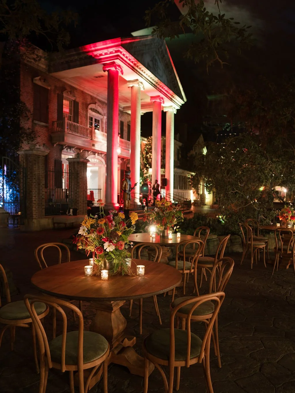 A lit-up outdoor patio with wooden tables and chairs, candles and flower arrangements, in front of a historic house with columns and a balcony, illuminated for an evening event with live music.