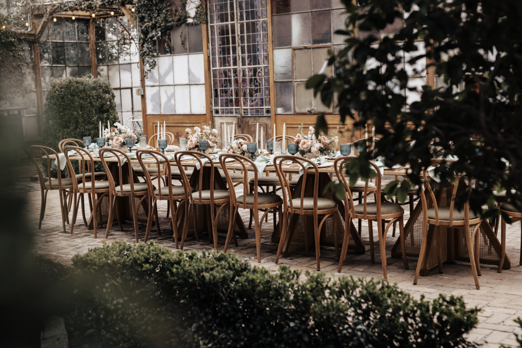A long outdoor dining table set with floral centerpieces, candles, and glassware, surrounded by wooden chairs, in a rustic garden setting with a greenhouse or shed in the background.
