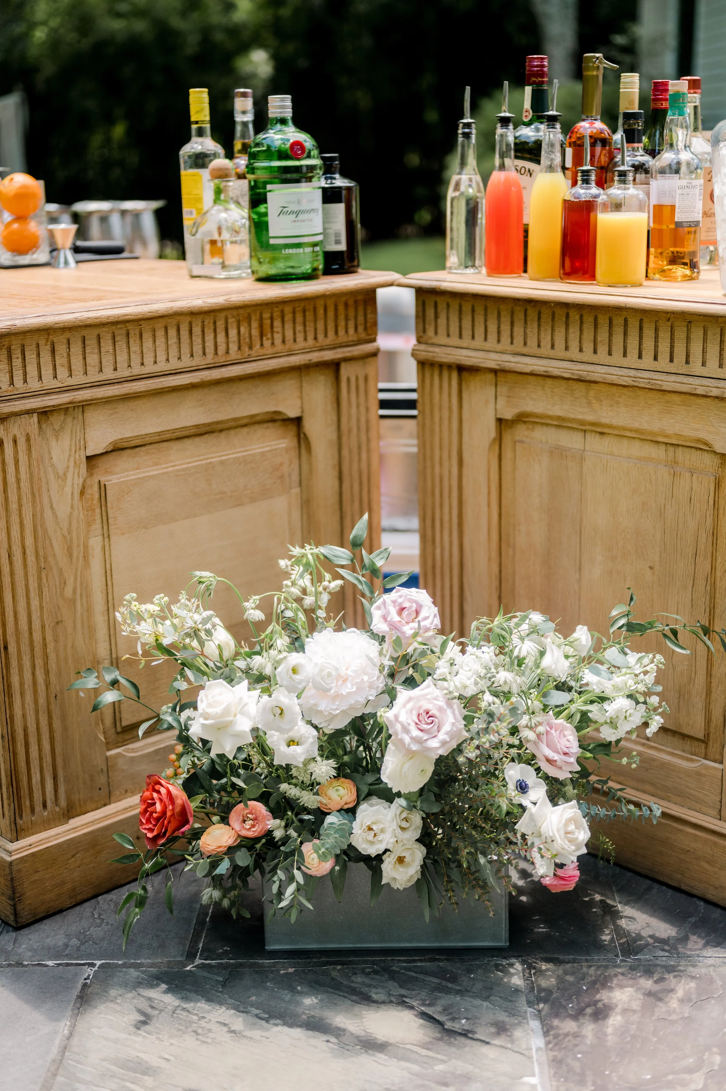 A large flower arrangement with white, pink, red, and peach flowers and green foliage placed in a gray rectangular container on a black tiled floor, with a wooden bar in the background.