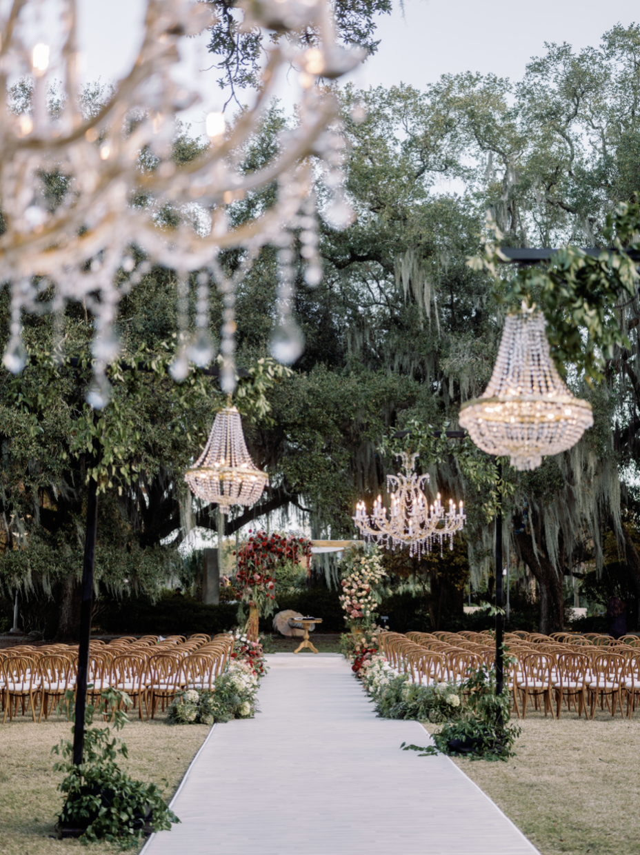 Outdoor wedding ceremony setup with rows of wooden chairs, floral arrangements, chandeliers hanging from trees, and a white aisle runner leading to an altar decorated with flowers.