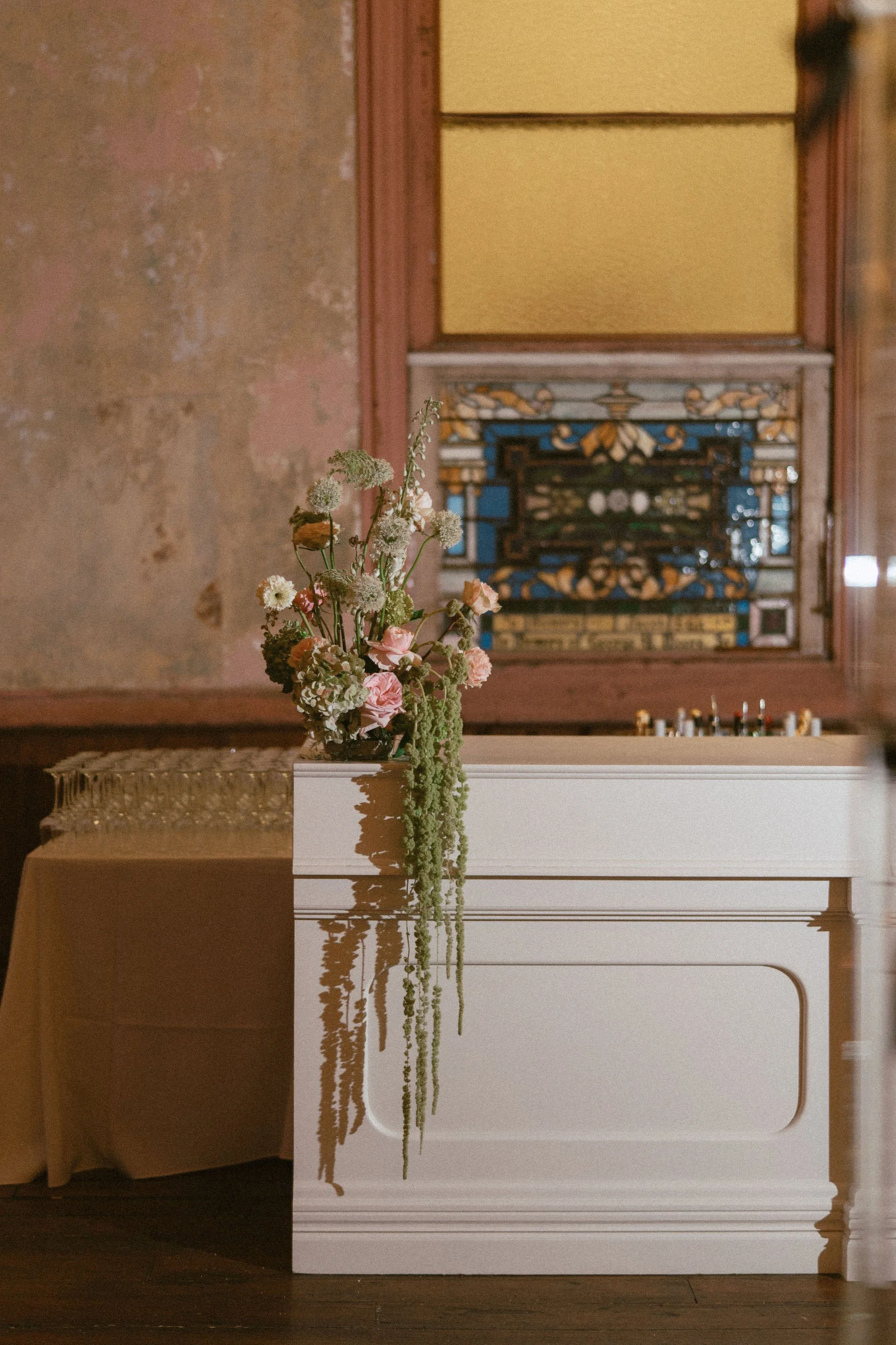 A bouquet of mixed flowers in a dark vase on a white table in a room with stained wall, yellow wall, and stained glass window.