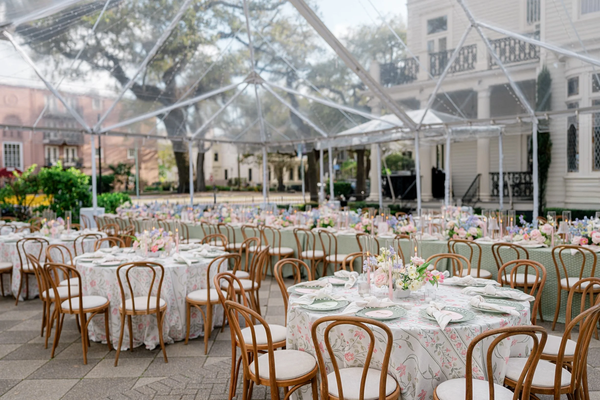 Outdoor wedding reception setup with decorated tables and chairs under a clear tent, surrounded by residential buildings and trees.