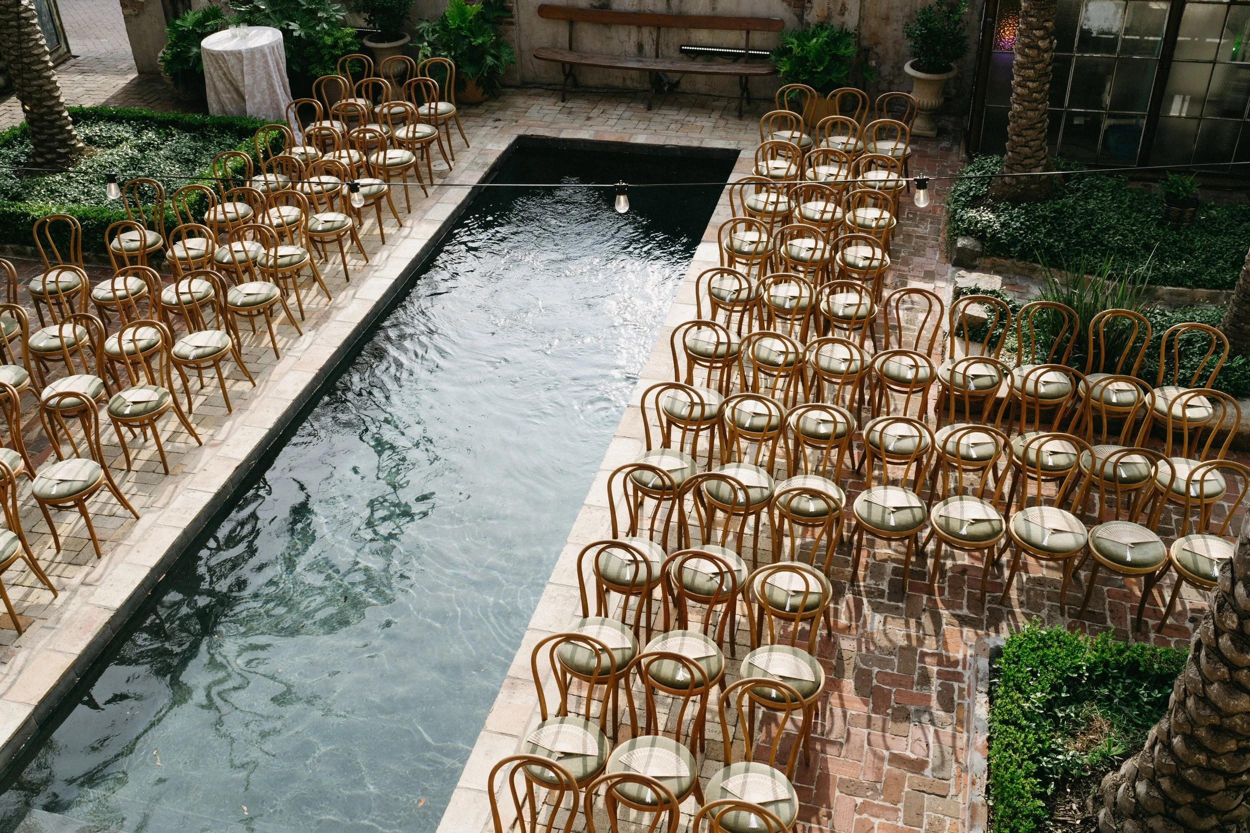 An indoor venue with a rectangular water feature in the center. Surrounding the water are numerous wooden chairs with cushioned seats arranged for an event. To the left, there is a tall white table with a tablecloth, and green plants in pots are plac