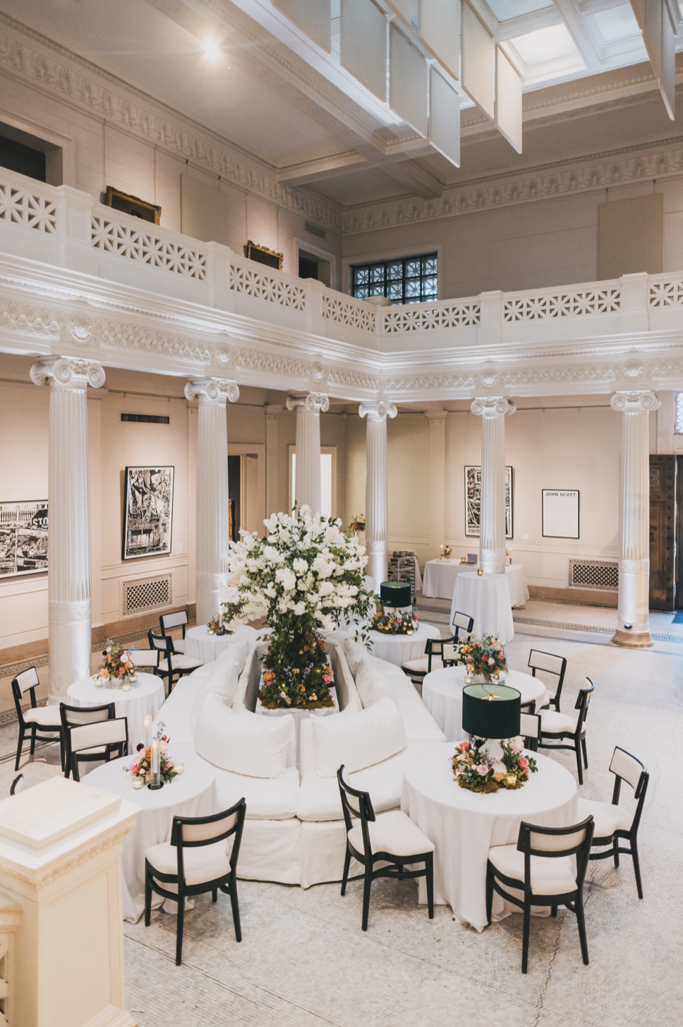Elegant event setup in a grand hall with white columns, a central large floral arrangement, and round tables with floral centerpieces and black and white chairs.