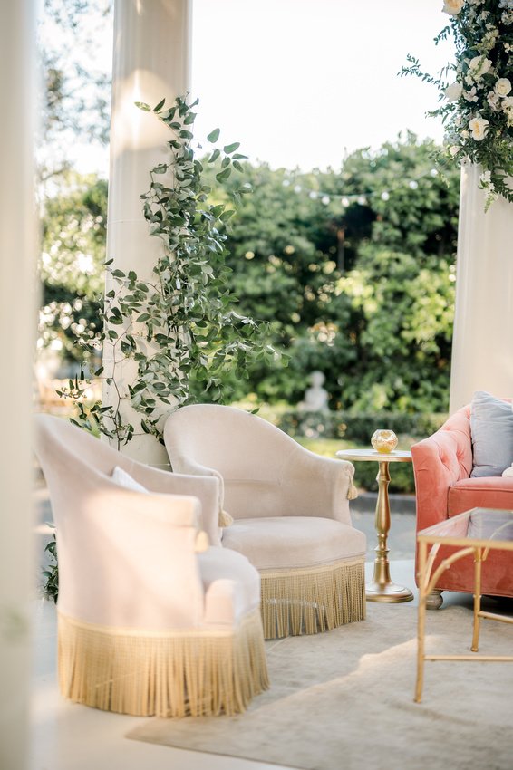 Elegant outdoor seating area with upholstered chairs, a pink sofa, a gold side table with a decorative object, and lush greenery in the background.