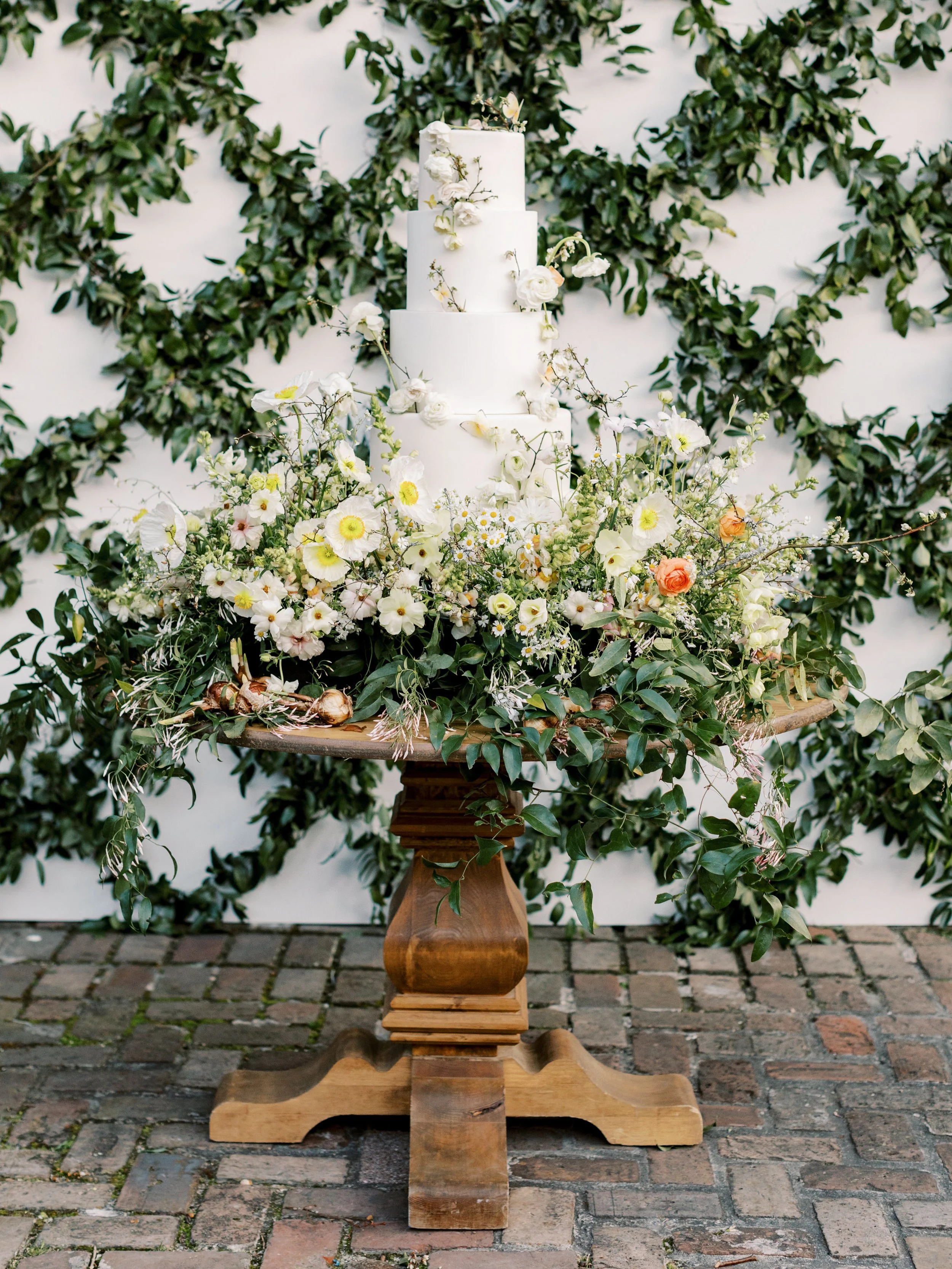 A tiered white wedding cake decorated with white flowers and greenery on a wooden stand, surrounded by white and peach flowers, against a backdrop of green leaves.
