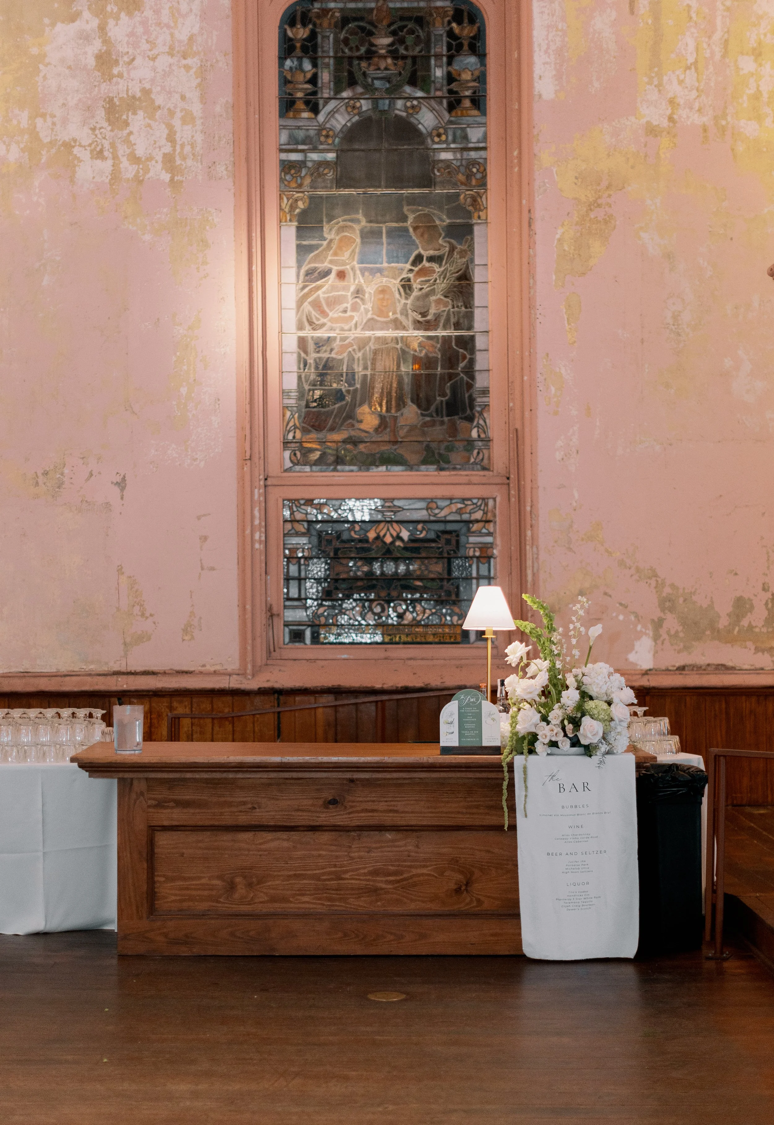 A bar counter with a bouquet of white flowers, a small table lamp, and a menu, set against a weathered pink wall with a stained glass window depicting a nativity scene.