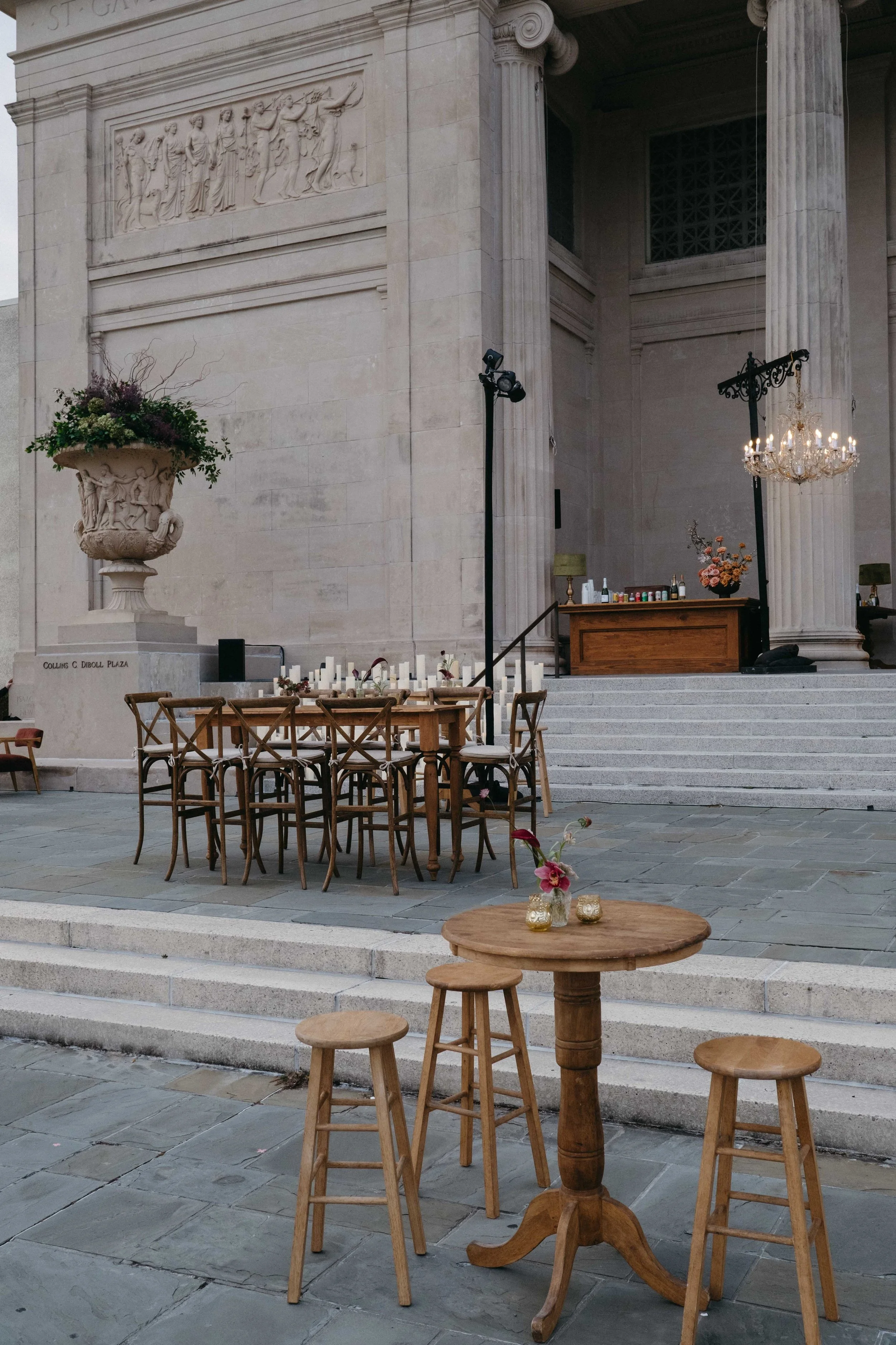 An outdoor event setup with a large stone building in the background, featuring columns, a chandelier, and a bas-relief sculpture. There are wooden tables and stools arranged for guests, with candles, flowers, and drinks on a wooden bar area.