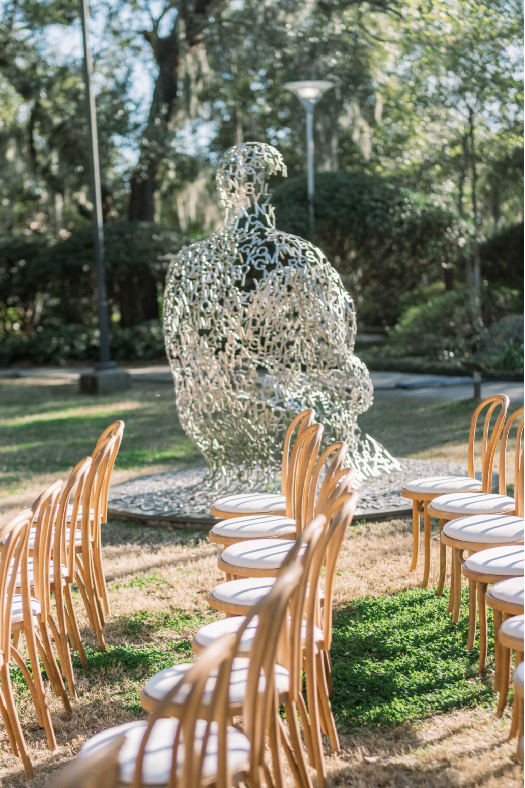 Outdoor art installation of a human head and shoulders sculpture made from interconnected metal letters, surrounded by chairs with white cushions arranged in a semi-circle in a park with trees and grass.