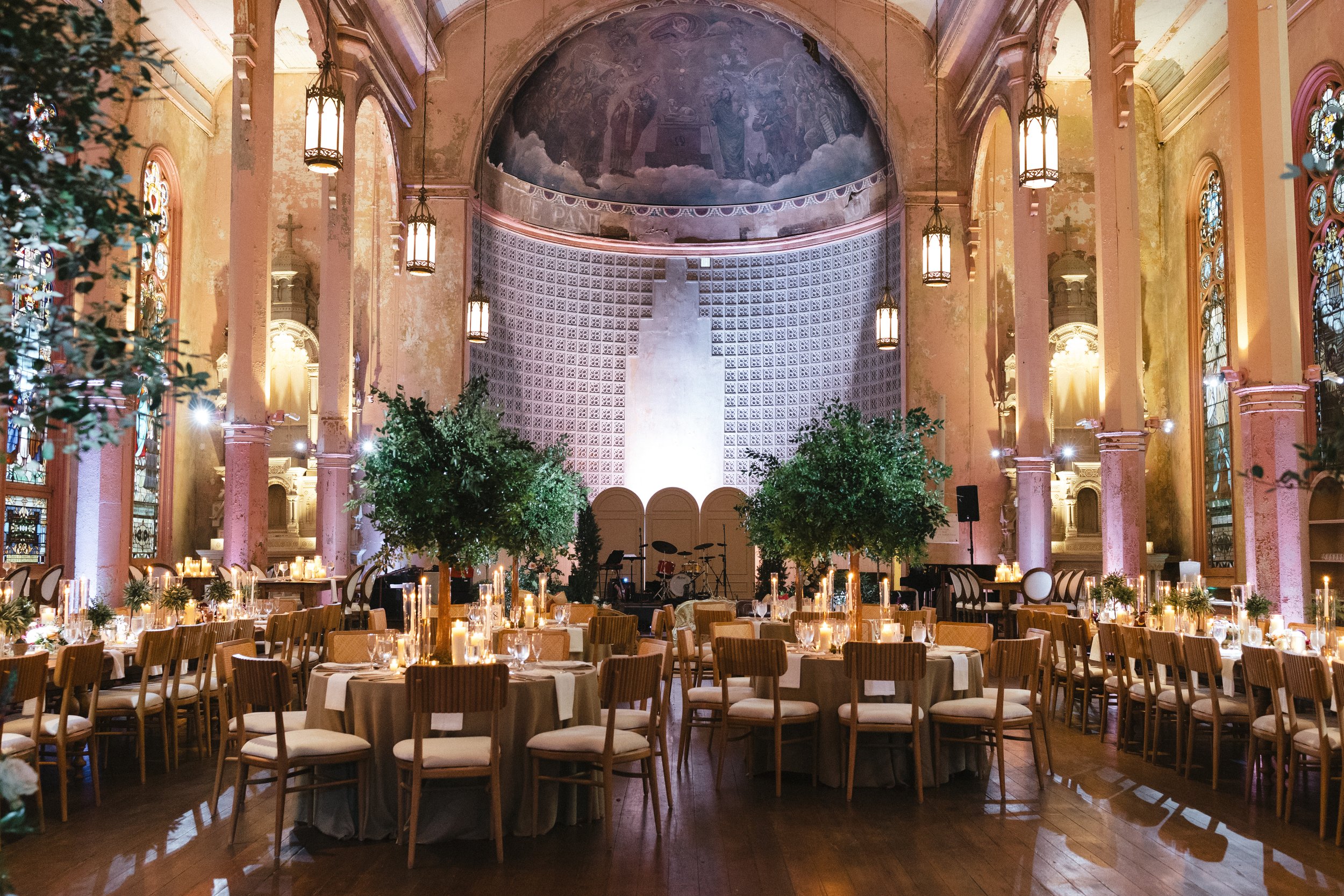 Inside a church decorated for a wedding reception with round tables, candles, and chairs, tall trees in the center, stained glass windows on the walls, and a large painting on the ceiling.