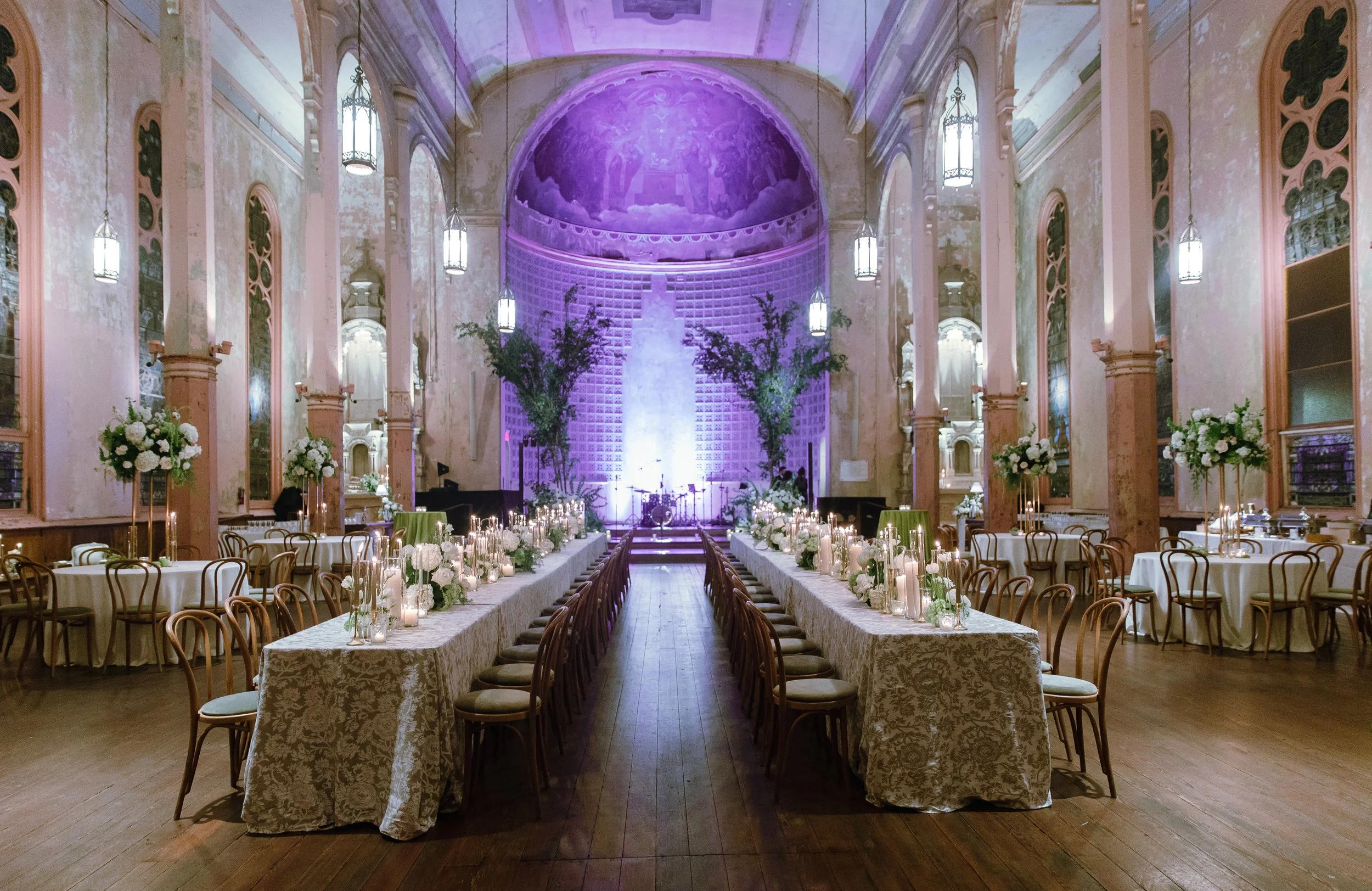 Elegant indoor event space with long banquet tables decorated with white tablecloths, floral centerpieces, and candles. The room features tall arches, chandeliers, and a purple-lit painted dome ceiling.