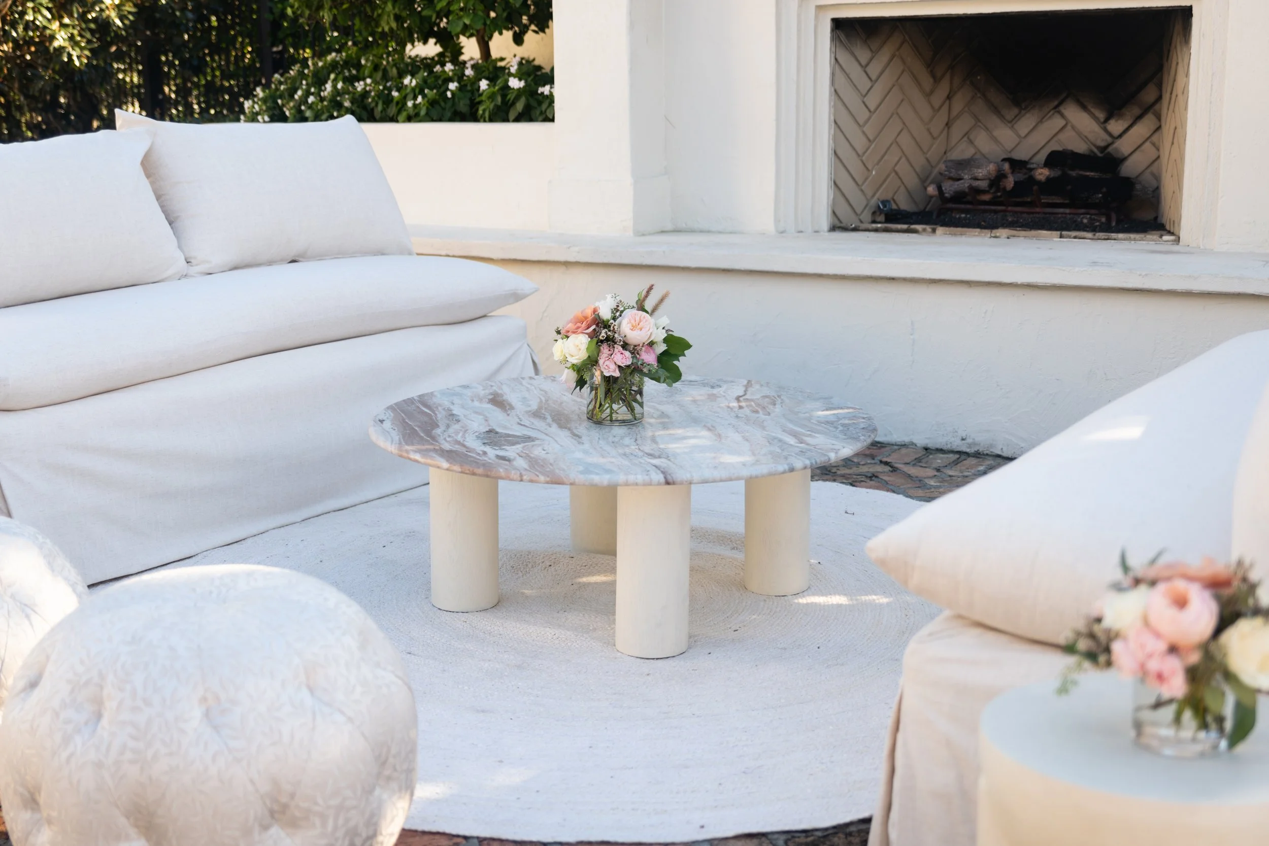 Outdoor patio with white seating and a round marble table holding a flower arrangement, in front of a fireplace with logs and a brick interior.