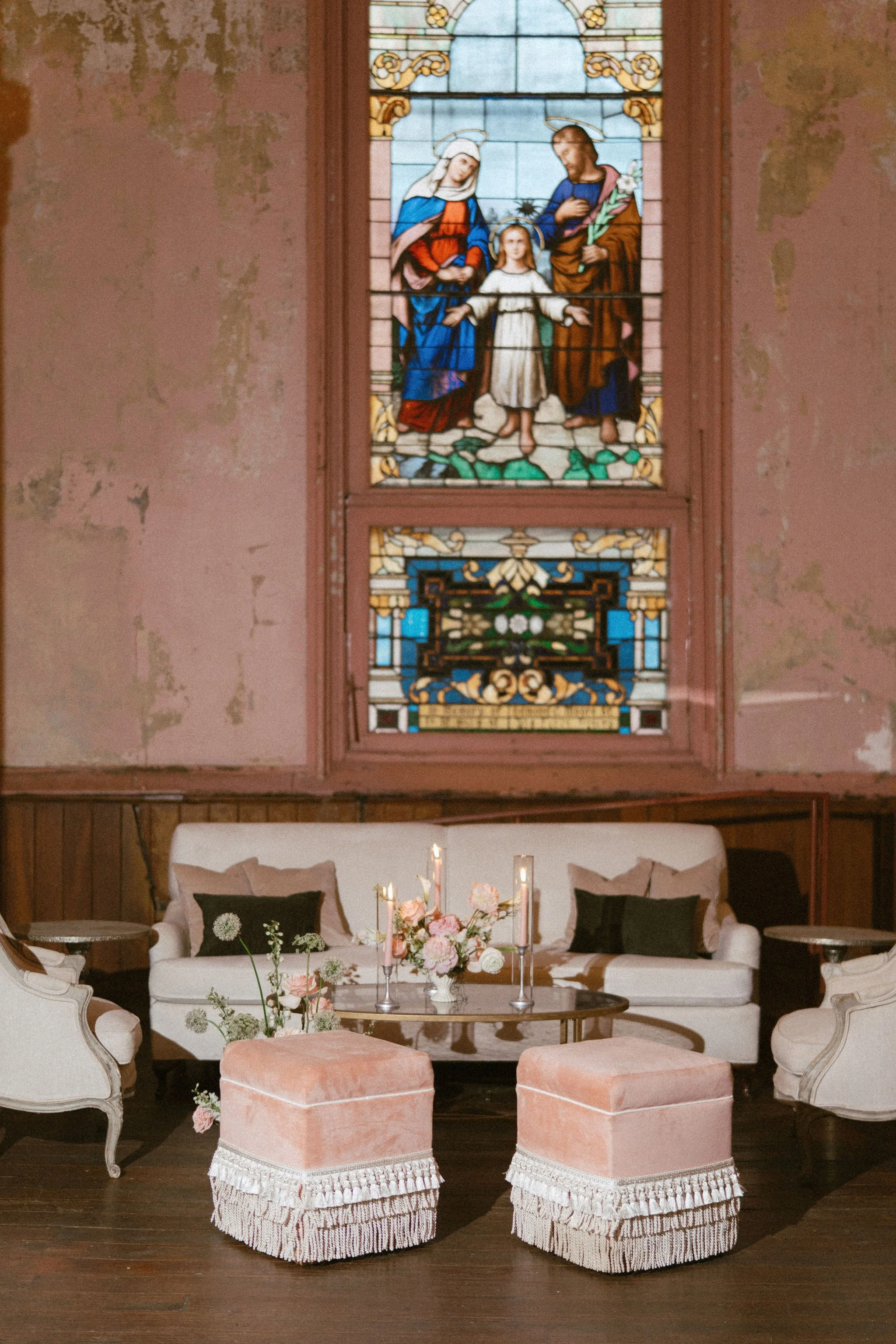A cozy living room with a white sofa adorned with black pillows, a floral centerpiece and pink velvet ottomans with fringe in front, and a stained glass window depicting religious figures hanging on a wall with peeling paint.