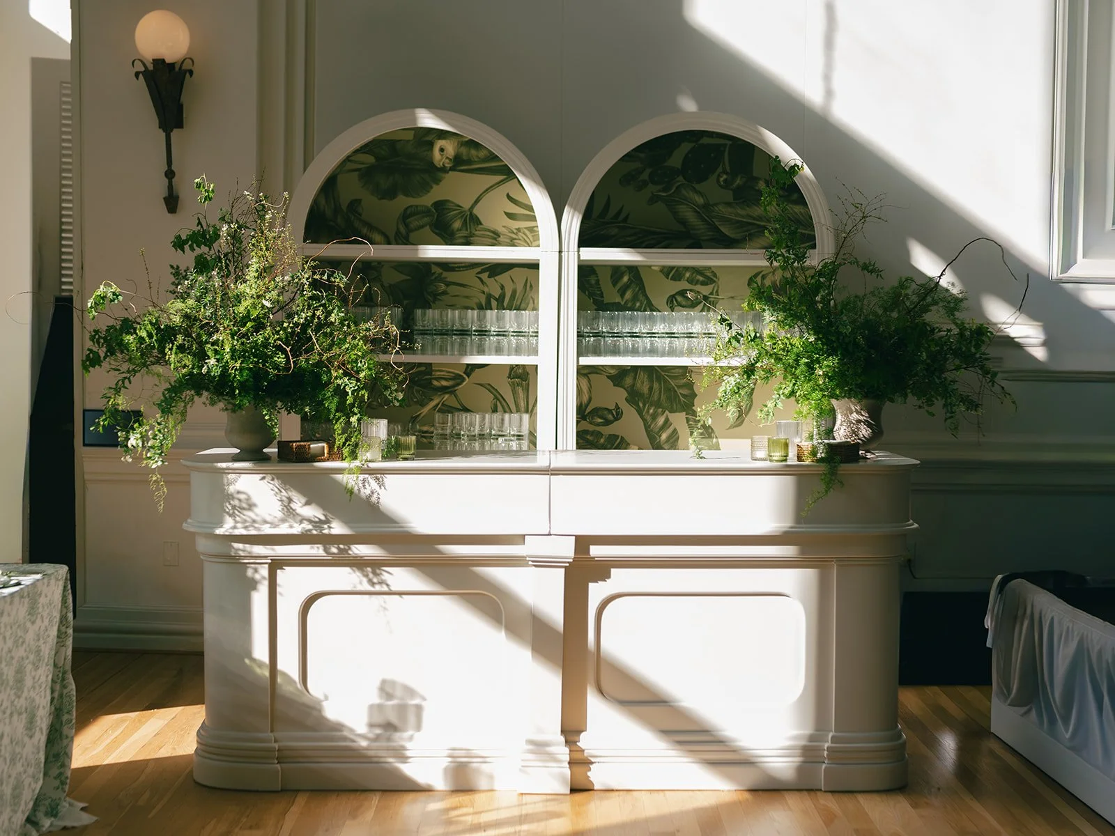 A white buffet table with large green plant arrangements on either side, set against a white wall with leafy patterned wallpaper. Glassware is arranged on the shelves behind the table, and sunlight casts shadows across the scene.