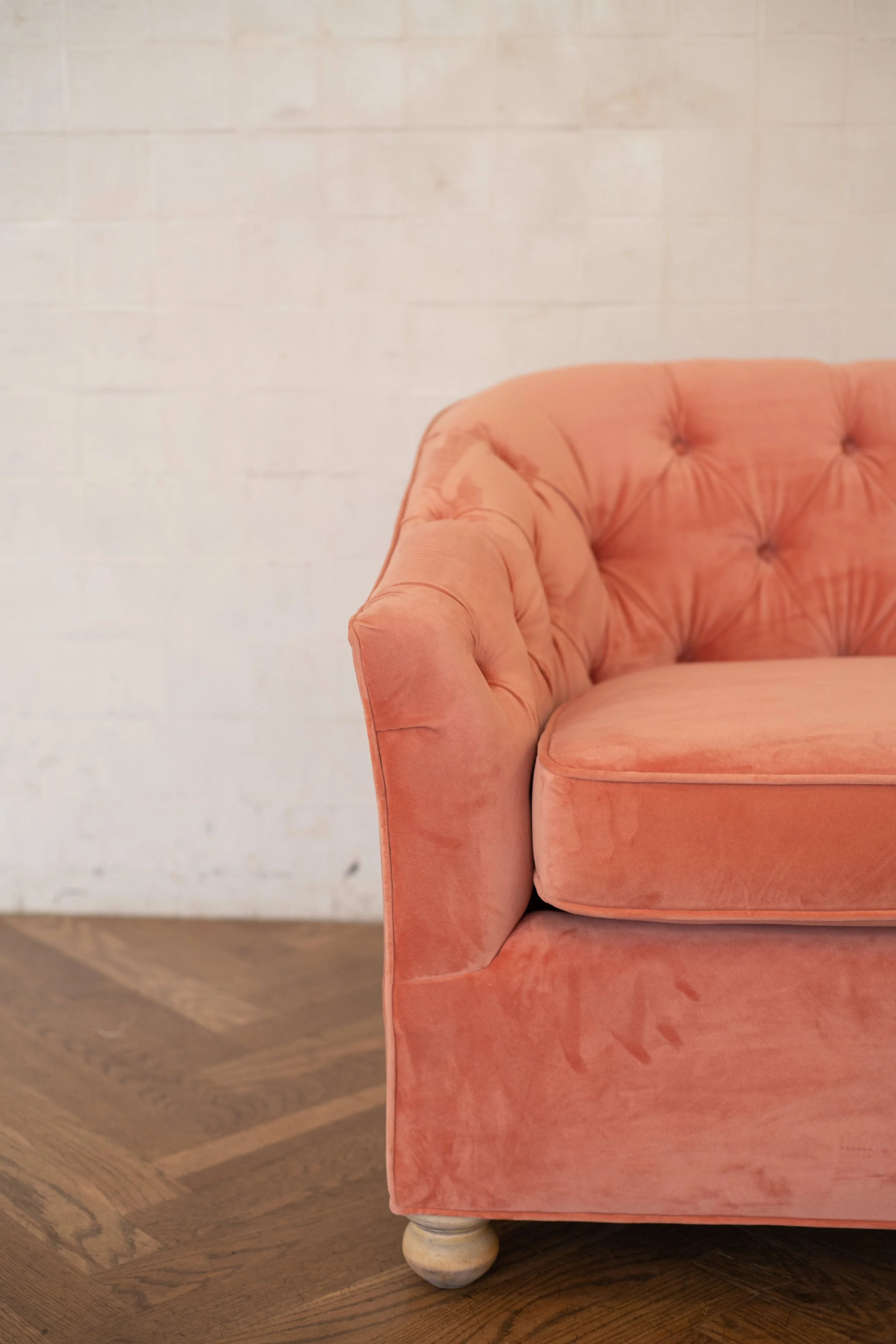 Close-up of an orange velvet sofa with tufted backrest, over a wooden floor with a white brick wall in the background.