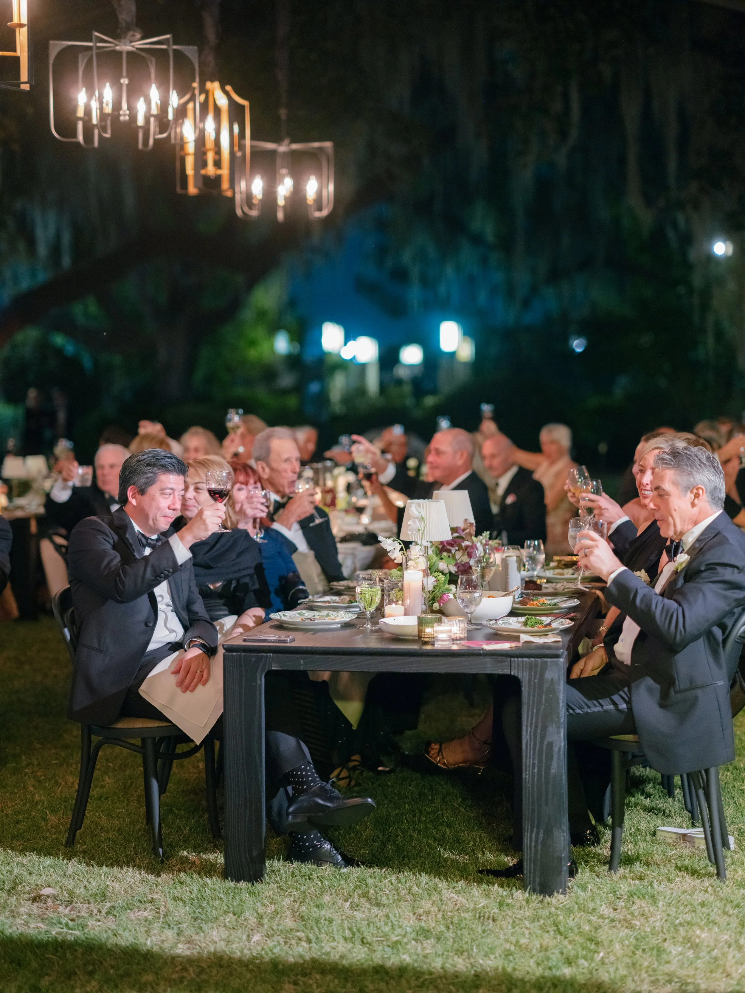 Elegant outdoor dinner party with guests in tuxedos and evening dresses raising glasses for a toast, under string lights at night.