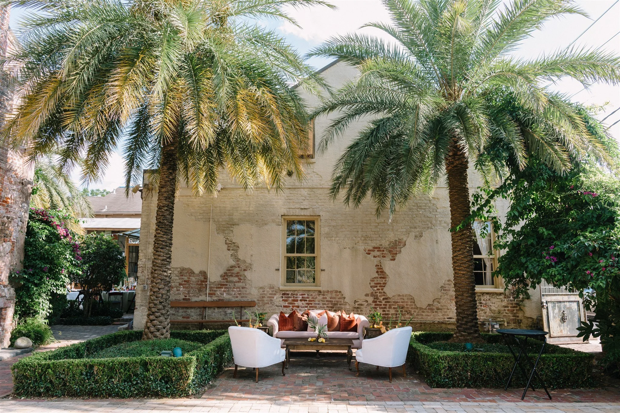 A backyard patio with a brick wall, two tall palm trees, a white vintage sofa with pink and white cushions, a small table with a potted plant, surrounded by green hedges and plants, and neighboring house walls and windows visible in the background.