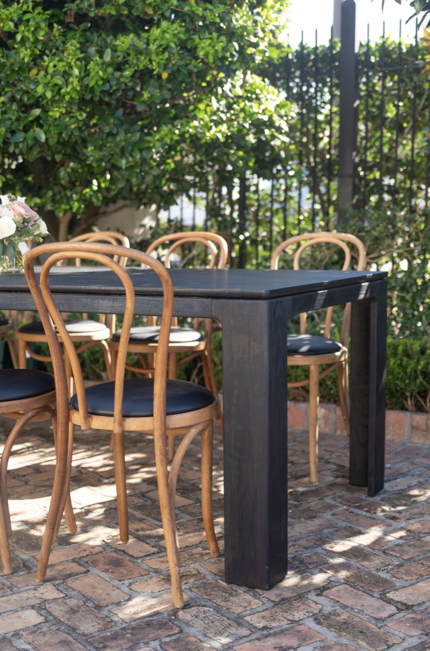 Outdoor patio table with wooden chairs, brick flooring, surrounded by lush greenery and bushes, with sunlight casting shadows.