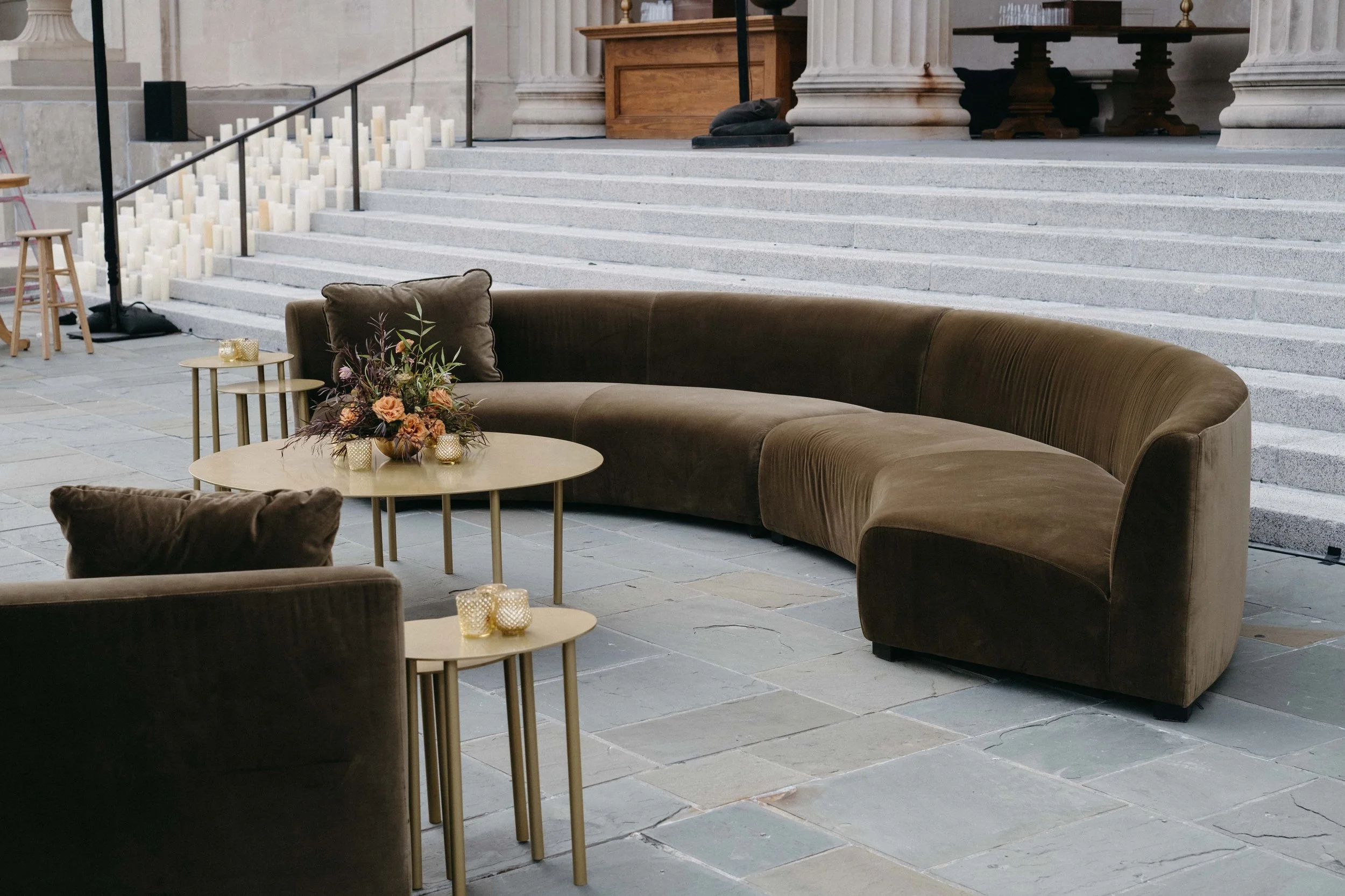 Interior of a building with stone steps, a brown curved sofa, a small round table with flower arrangement and candles, and scattered candles, with some items like a ladder and black bag nearby.