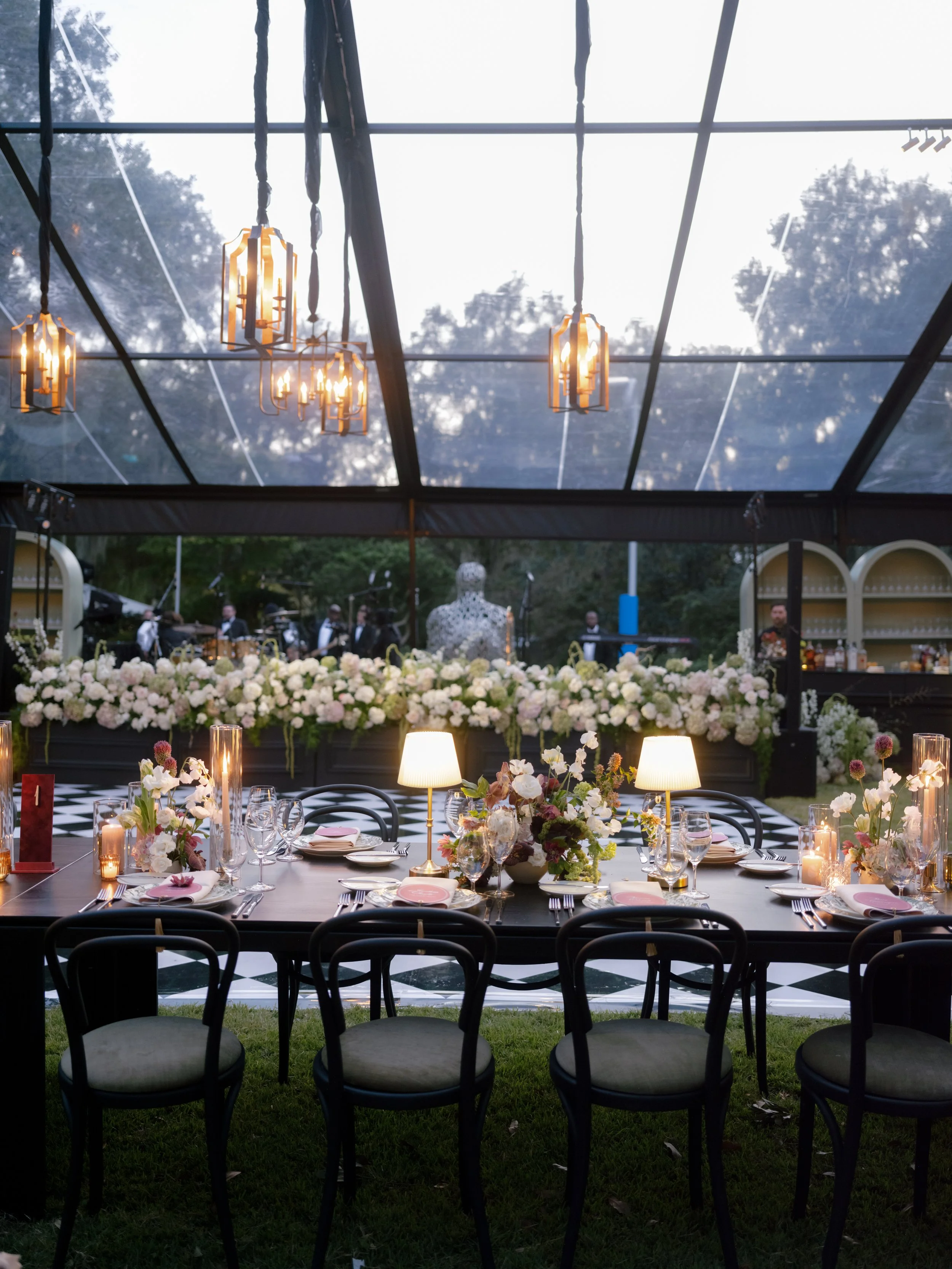 Elegant indoor-outdoor dining setup with round table, floral centerpieces, candles, and plates. Overlooks a garden with trees and a decorative mannequin statue in the background. Black and white checkered floor adds a stylish touch.