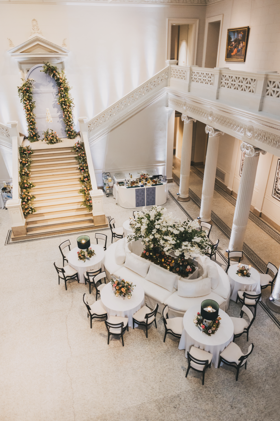 Elegant indoor event space with a grand staircase decorated with floral arrangements, a central seating area with a large white sofa surrounded by small round tables with floral centerpieces, and a bar area against the wall. The space has tall pillar