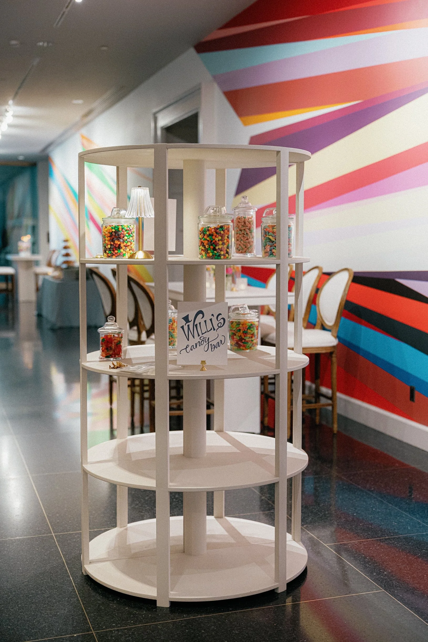 Empty white display shelf with jars of colorful candies in a decorated event space with a rainbow-striped wall.