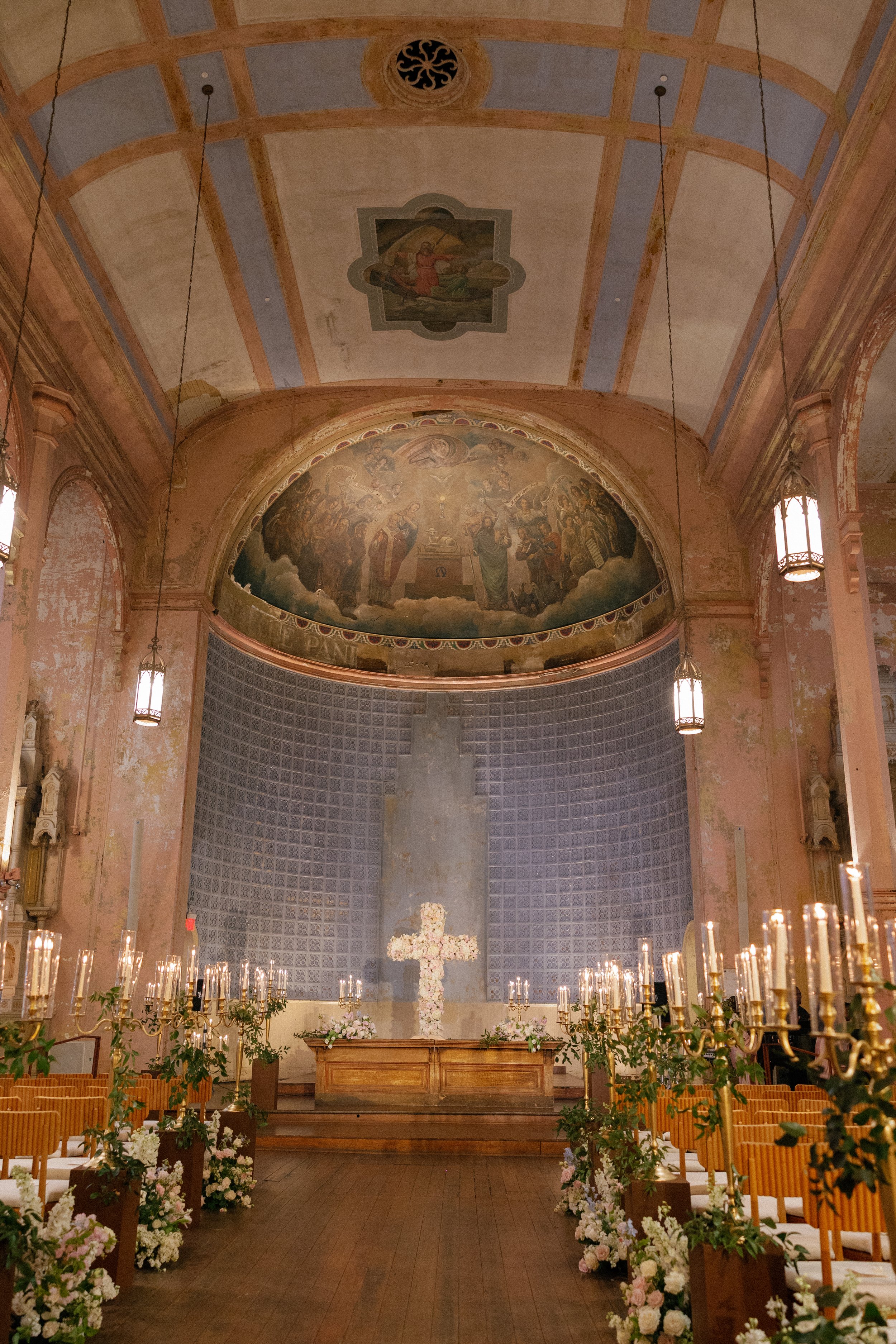 Interior of a church or chapel decorated for a wedding, with flower arrangements and candle holders along the aisle, and a cross at the altar. A large mural depicting a religious scene is on the wall behind the altar. The ceiling is painted with fram
