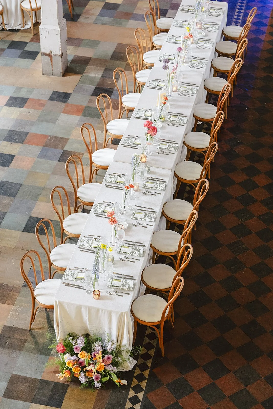 Long banquet table set for a celebration with floral centerpieces, candles, and place settings. Semi-circle wooden chairs surround the table, which is covered with a white tablecloth and decorated with colorful flowers at the end.