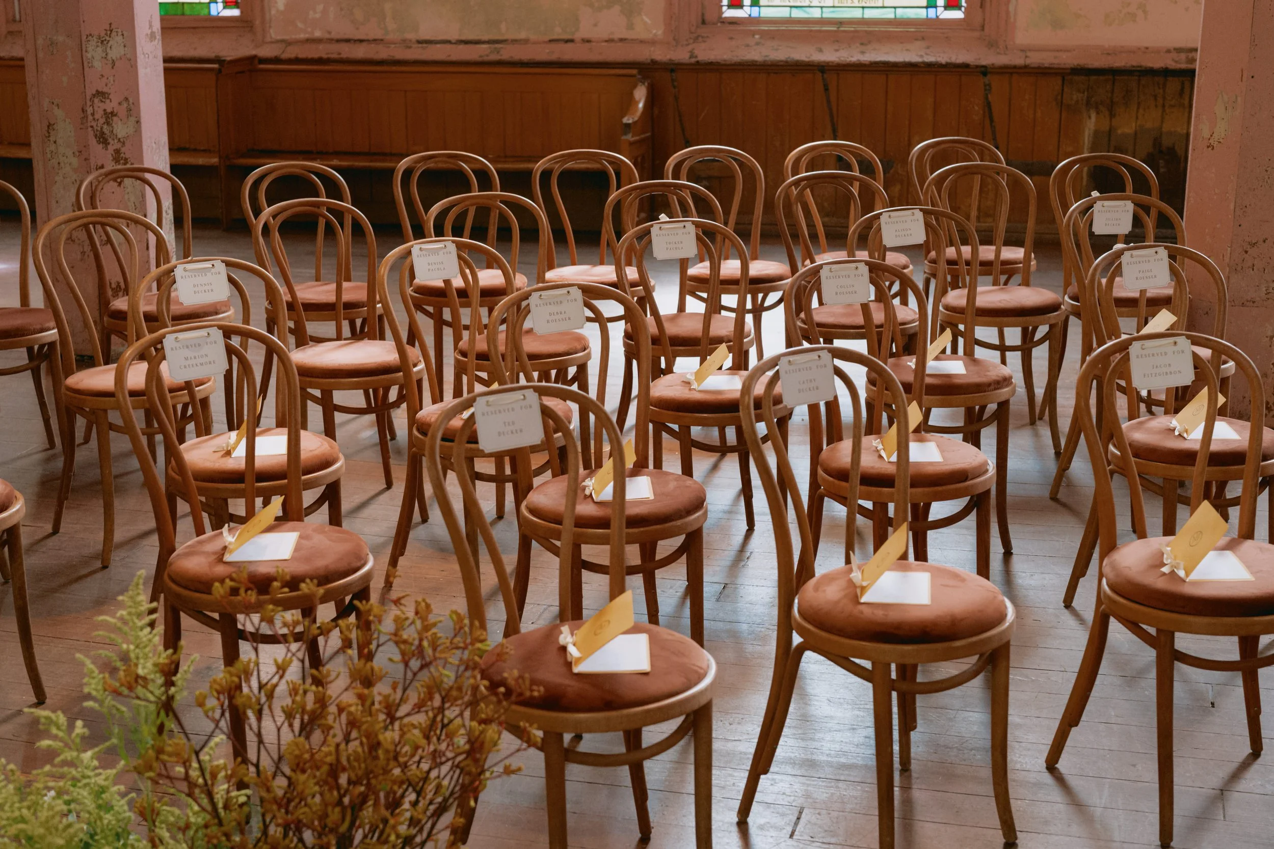 Multiple wooden chairs with beige cushions arranged in rows inside a rustic room with stained glass windows. Each chair has a reservation card on it.