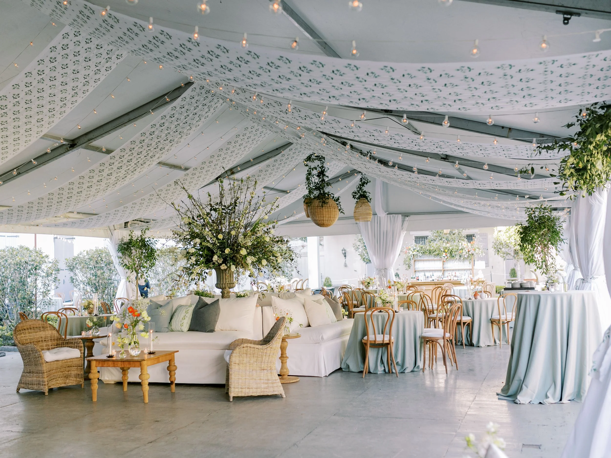 Interior of a decorated wedding or event venue with hanging string lights, floral arrangements, wicker and wooden chairs, and draped fabric on the ceiling.
