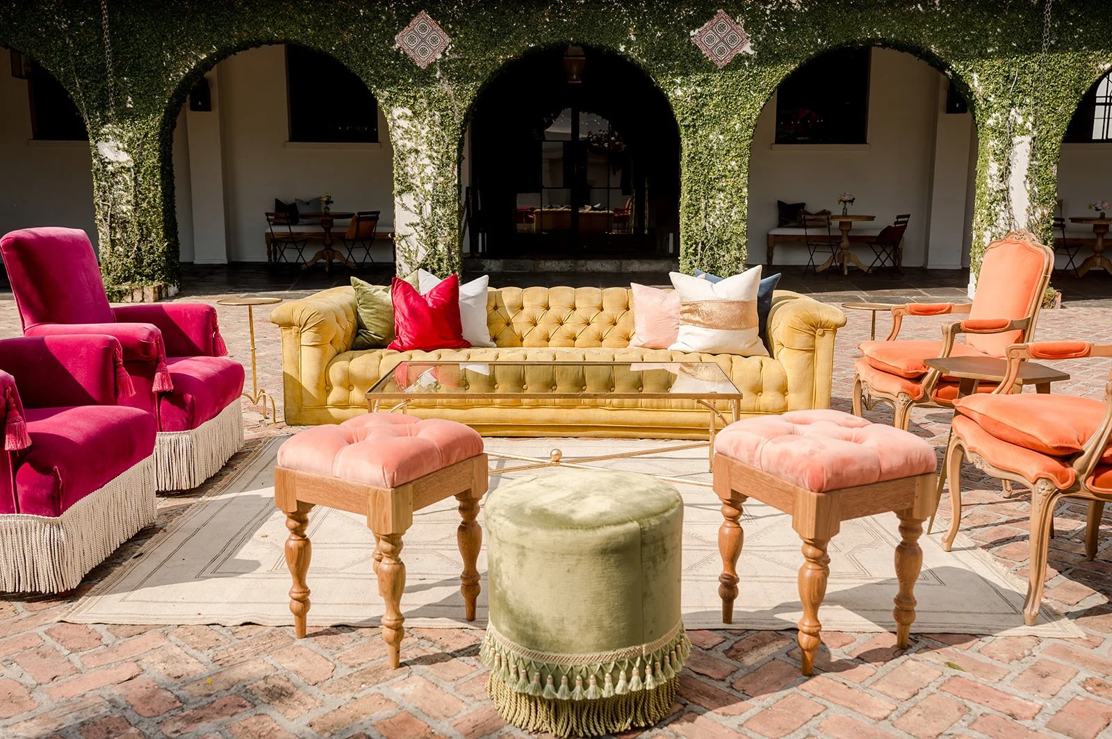 Colorful outdoor patio seating area with vintage style chairs, cushioned stools, a tufted sofa with pillows, and a glass coffee table, set against a backdrop of green ivy-covered arches.