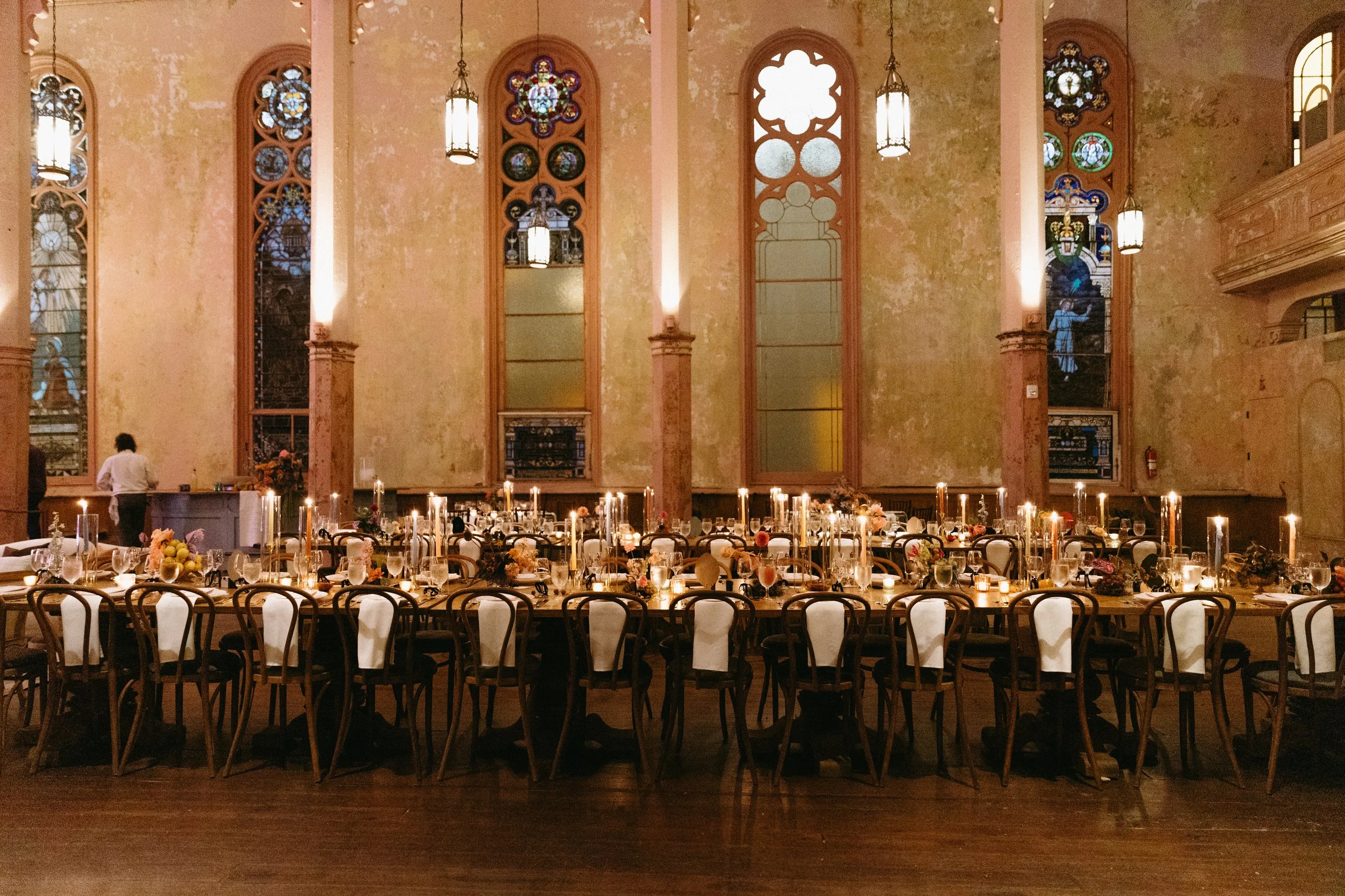 Long dining table set with candles, flowers, and place settings in a historic church or hall with stained glass windows, tall ornate wall sconces, and a high, textured ceiling.
