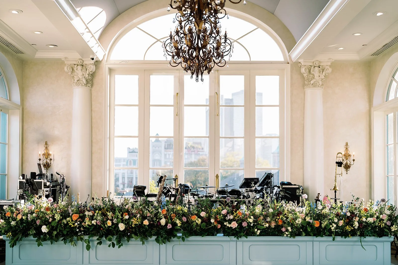 Elegant event stage with floral arrangements, musical instruments, and sound equipment in front of large, arched windows with a view of buildings outside. A chandelier hangs from the ceiling, and gold accents adorn the wall sconces and window handles